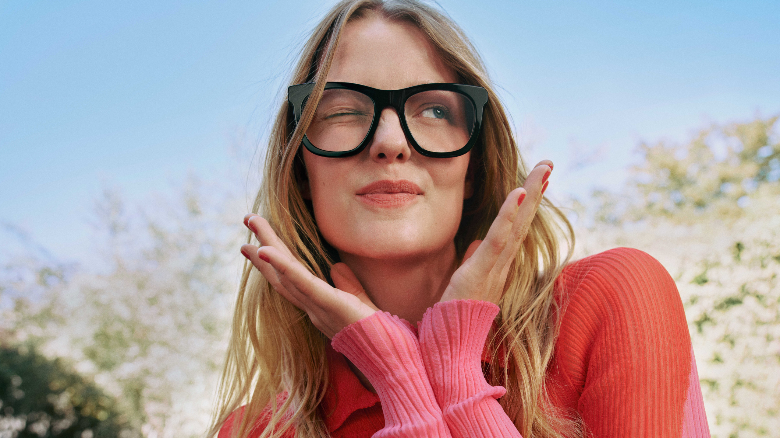 Una donna con lunghi capelli biondi, grandi occhiali neri e un maglione rosa fa l’occhiolino in modo giocoso, con le mani sotto il mento, davanti a uno sfondo esterno sfocato.