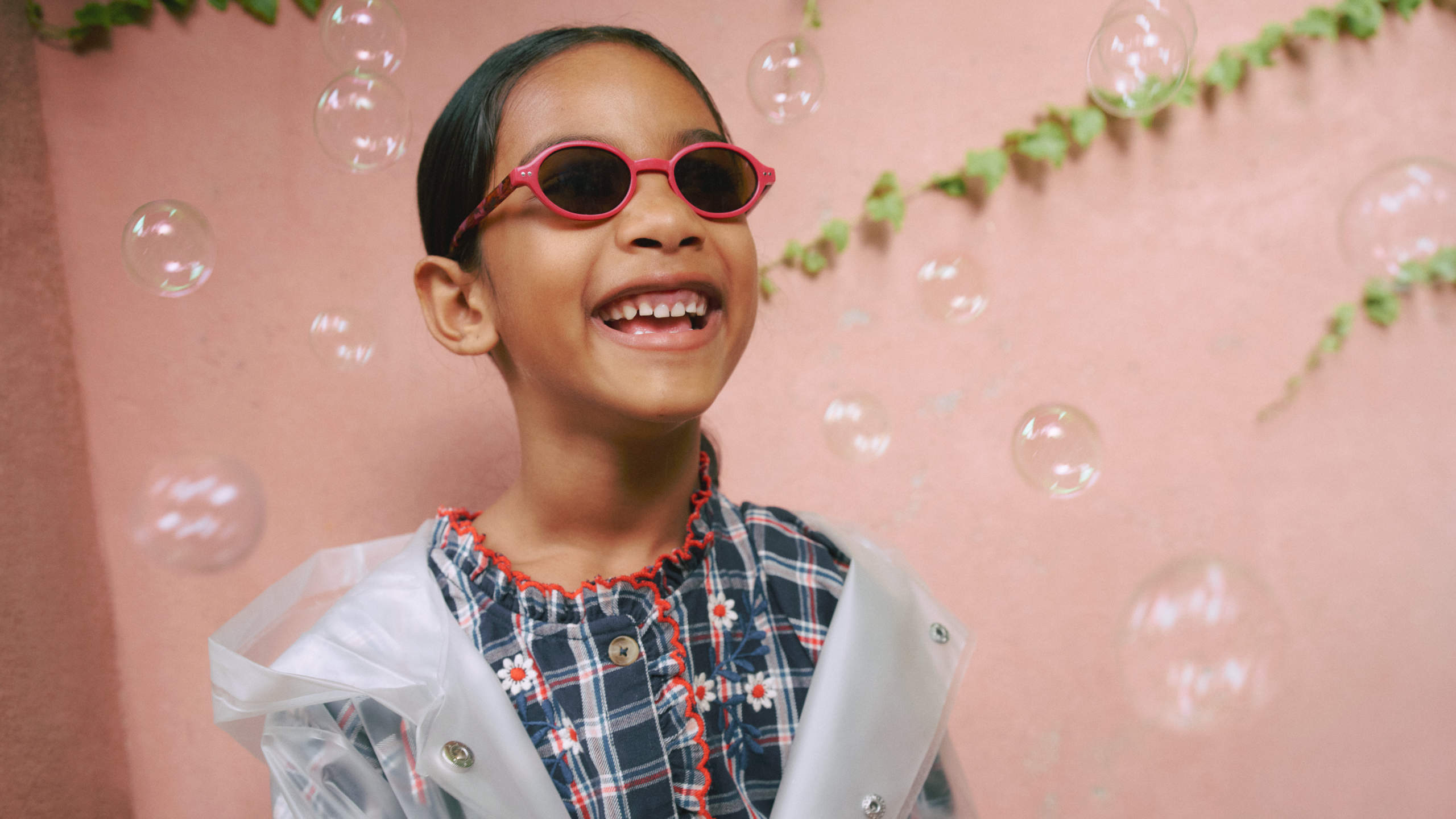 Un bambino sorridente con occhiali da sole rossi e una camicia a quadri, circondato da bolle di sapone, è in piedi davanti a una parete rosa con foglie verdi.