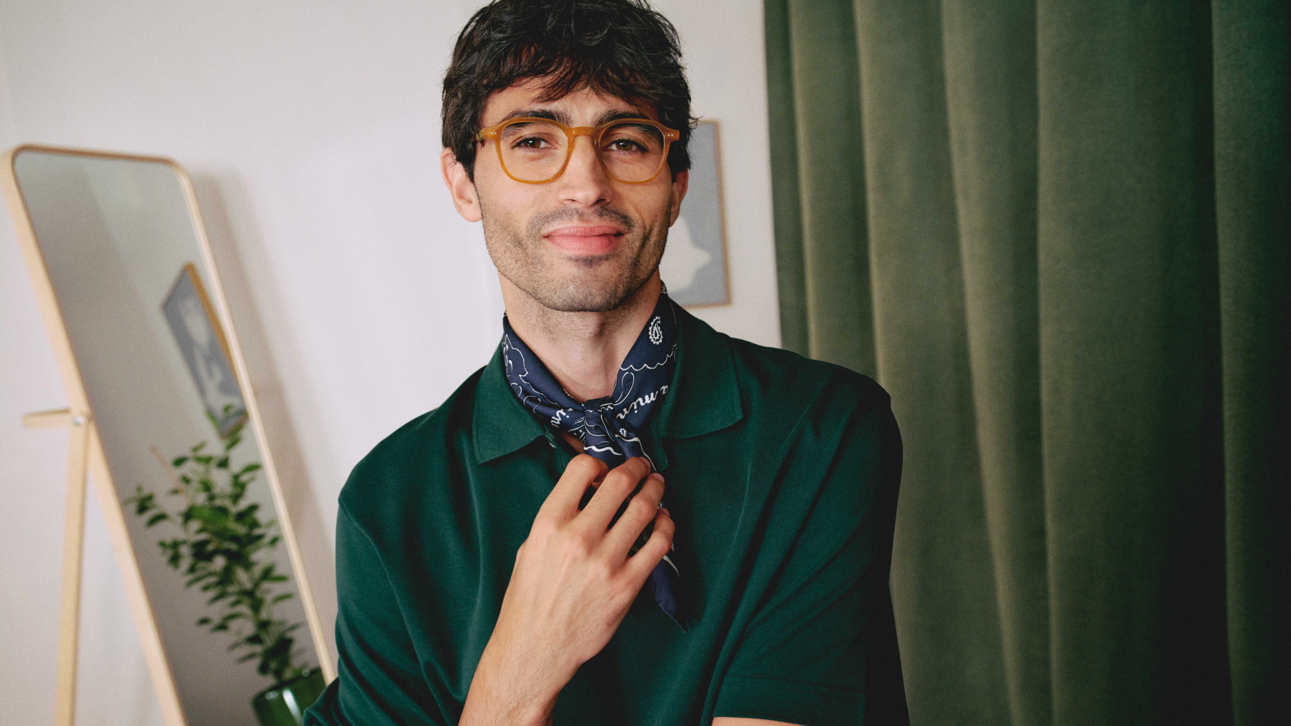 Ein Mann mit Brille und Schal lächelt leicht und steht in einem Raum mit einem grünen Vorhang und einem Spiegel.