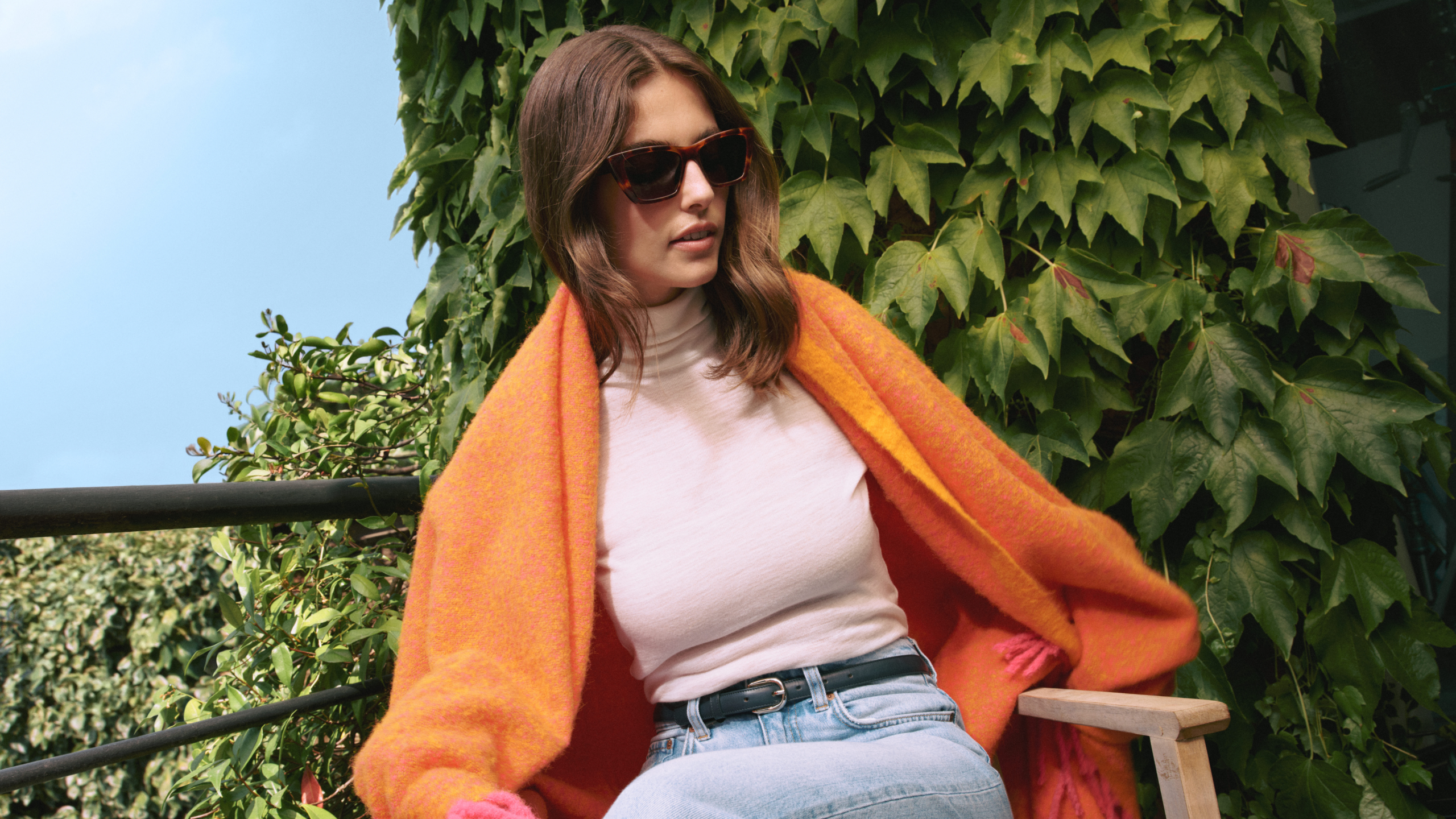 Eine Frau mit Sonnenbrille, weißem Rollkragenpullover, Jeans und einem orangefarbenen Schal sitzt auf einem Holzstuhl vor einem Hintergrund aus grünem Efeu.