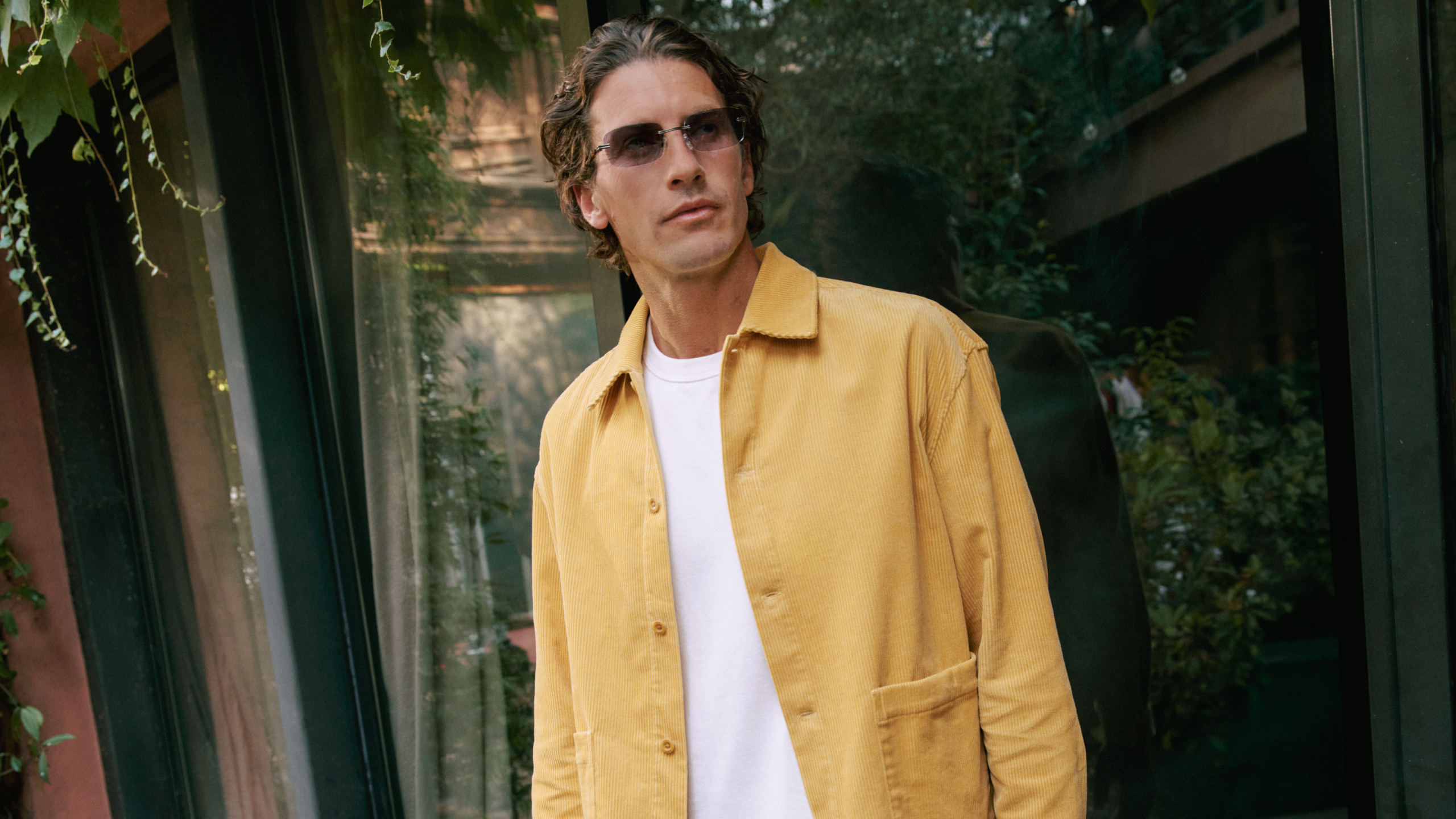 Ein Mann mit Sonnenbrille und gelber Jacke steht vor einer Glaswand, hinter ihm spiegelt sich Grün.