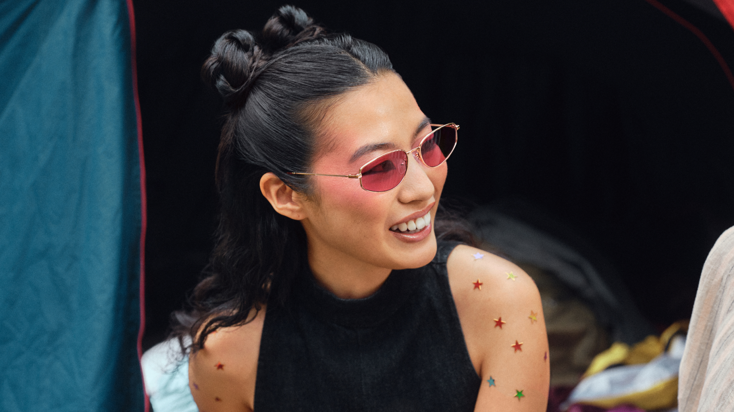 Eine Frau mit Sternenaufklebern auf den Armen, die eine rosa Sonnenbrille und ein schwarzes Oberteil trägt, lächelt, während sie neben einem blauen Zelt sitzt.