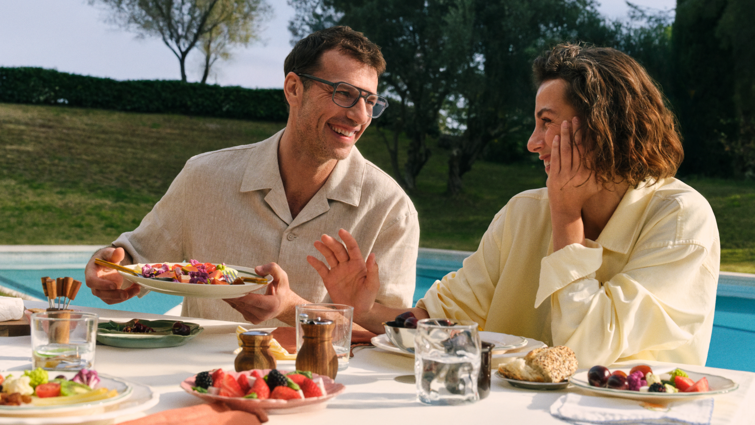 Zwei Menschen, die lächelnd an einem Tisch im Freien am Pool sitzen und gemeinsam essen, umgeben von verschiedenen Speisen und Getränken, mit Grünpflanzen im Hintergrund.