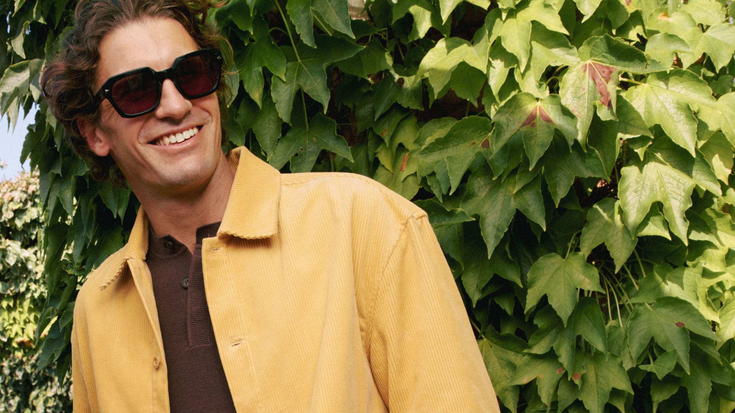 Ein Mann mit Sonnenbrille und gelber Jacke lächelt, während er vor einem grünen, dicht belaubten Hintergrund steht.