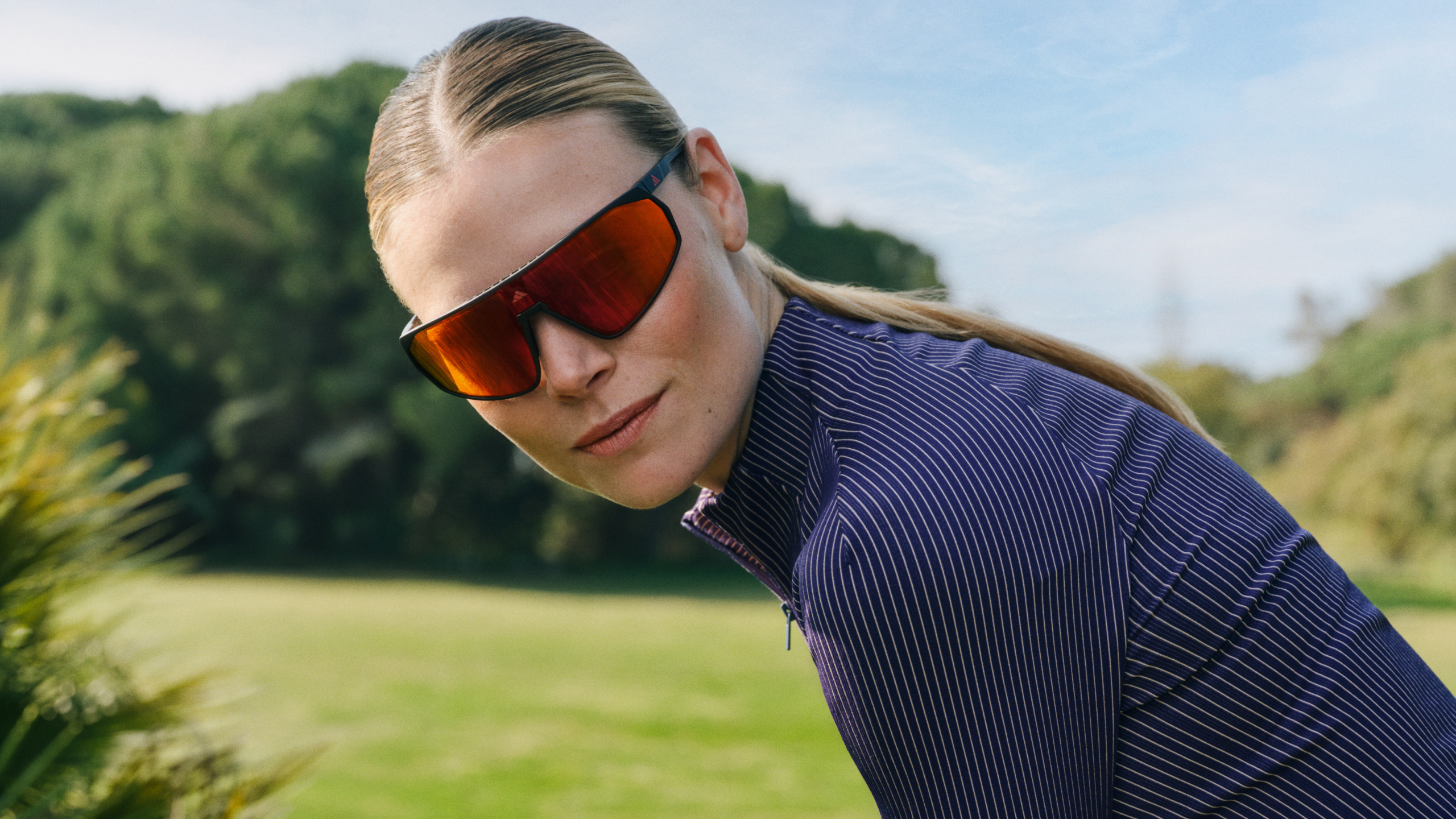 Eine Person mit einer großen roten Sonnenbrille und einer gestreiften Jacke, die im Freien vor einem grünen Hintergrund steht.