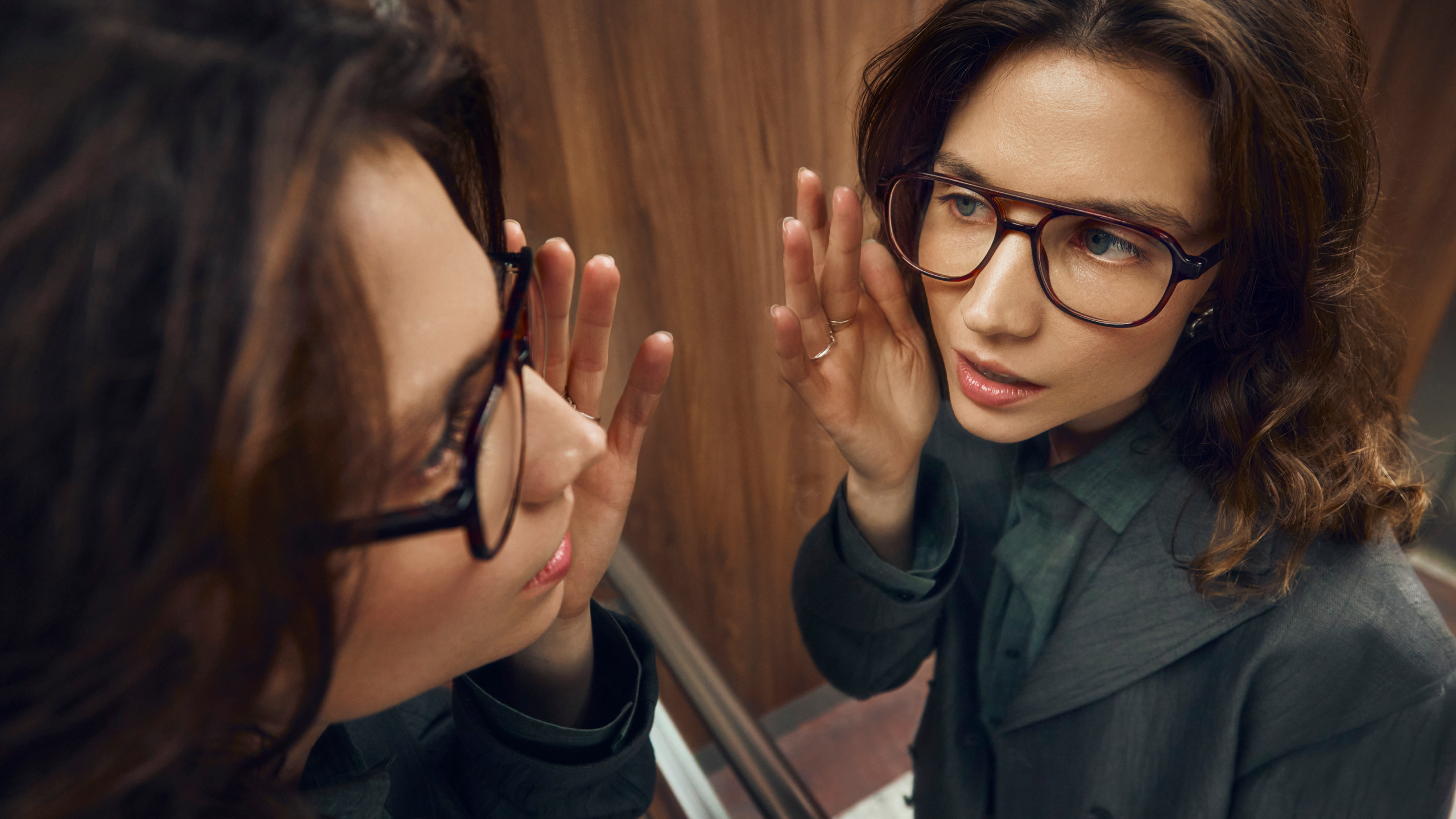 Une femme portant des lunettes et un blazer observe attentivement son reflet dans le miroir, la main près du visage.