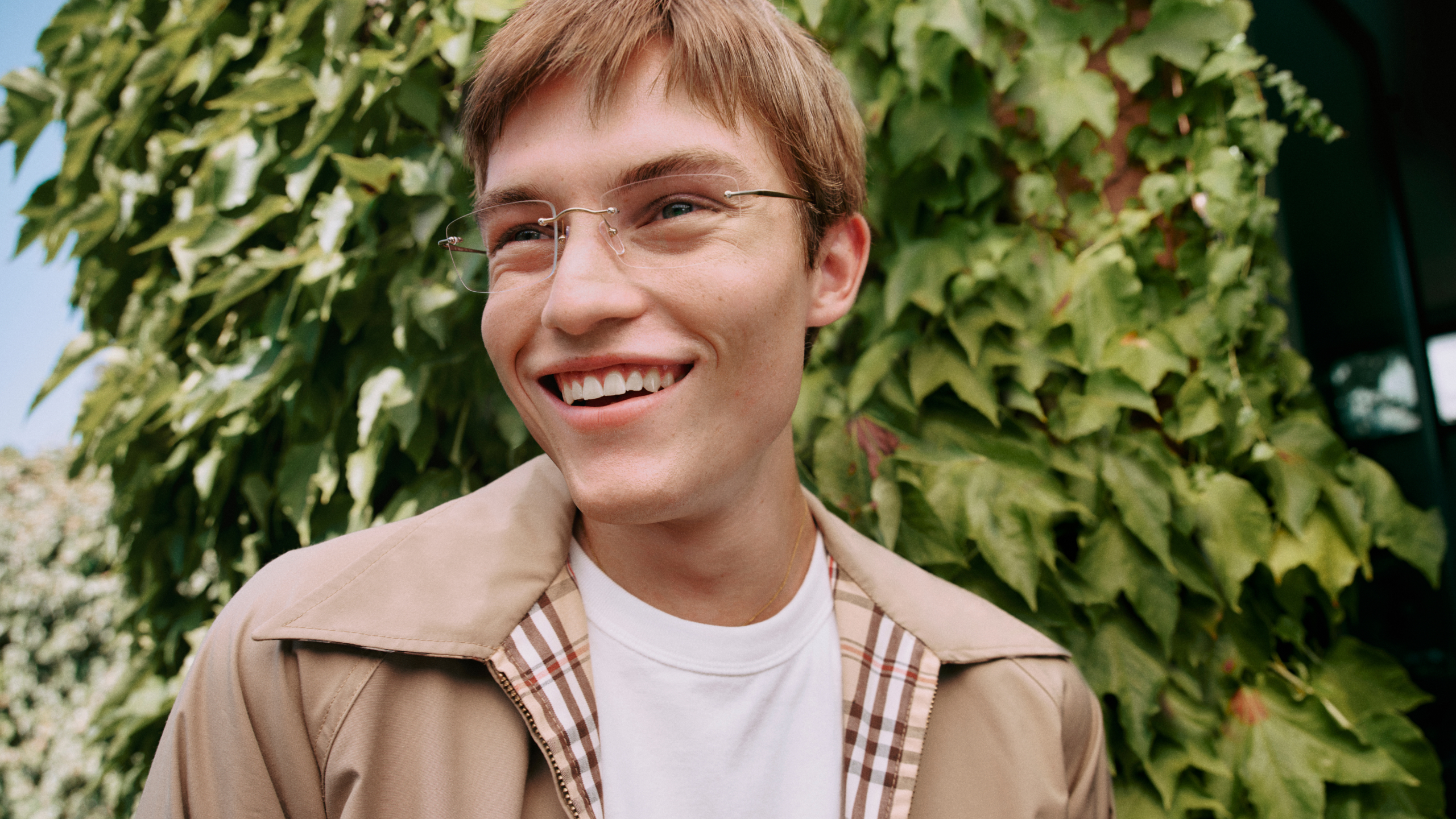 Eine lächelnde junge Person mit Brille und kurzen Haaren steht vor einer üppigen, grünen, mit Efeu bewachsenen Wand und trägt eine beige Jacke über einem weißen Hemd.