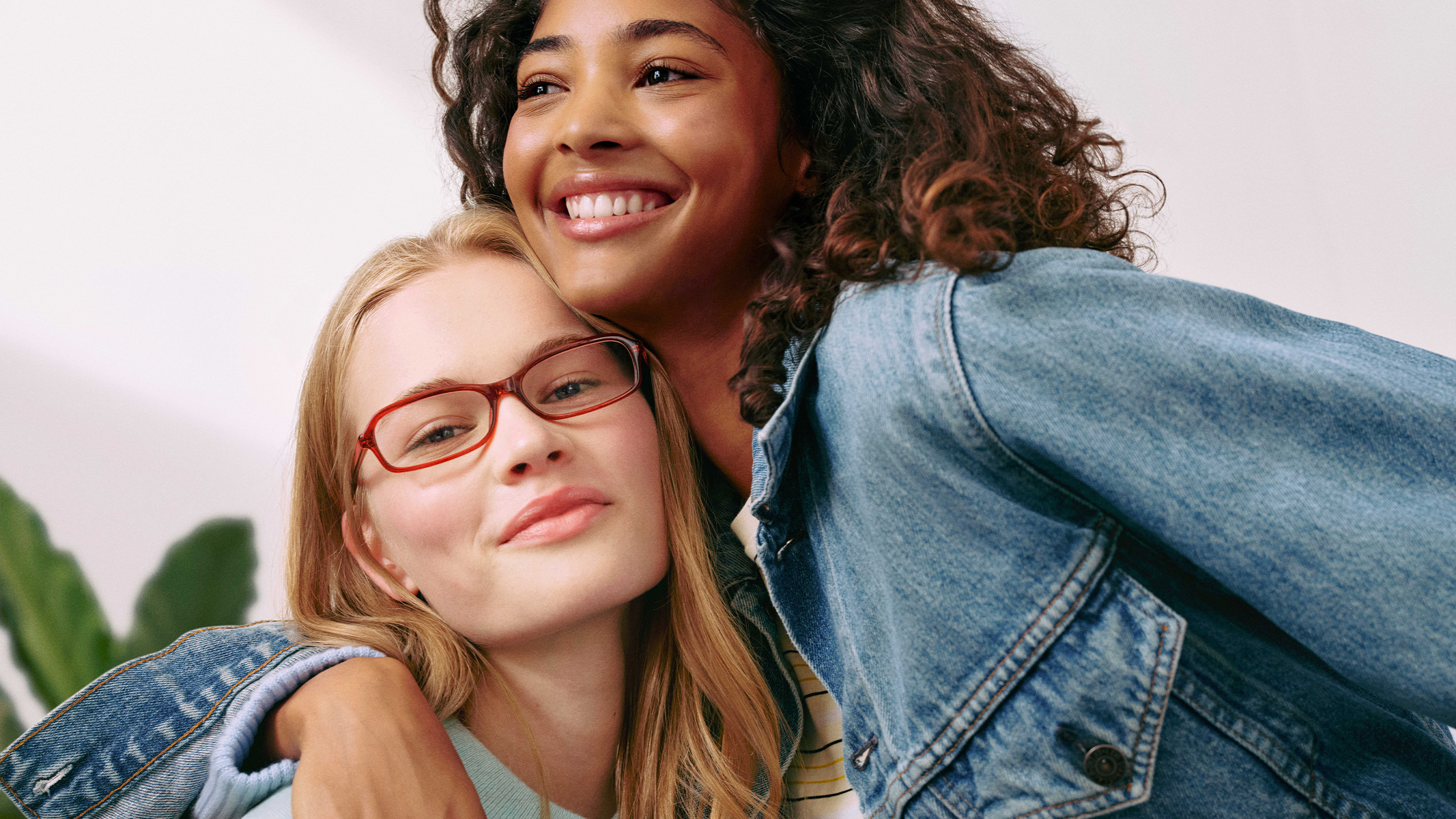Due donne che sorridono insieme, una delle quali indossa gli occhiali; assumono una posa rilassata e indossano giacche di jeans, davanti a uno sfondo chiaro con una pianta sullo sfondo.