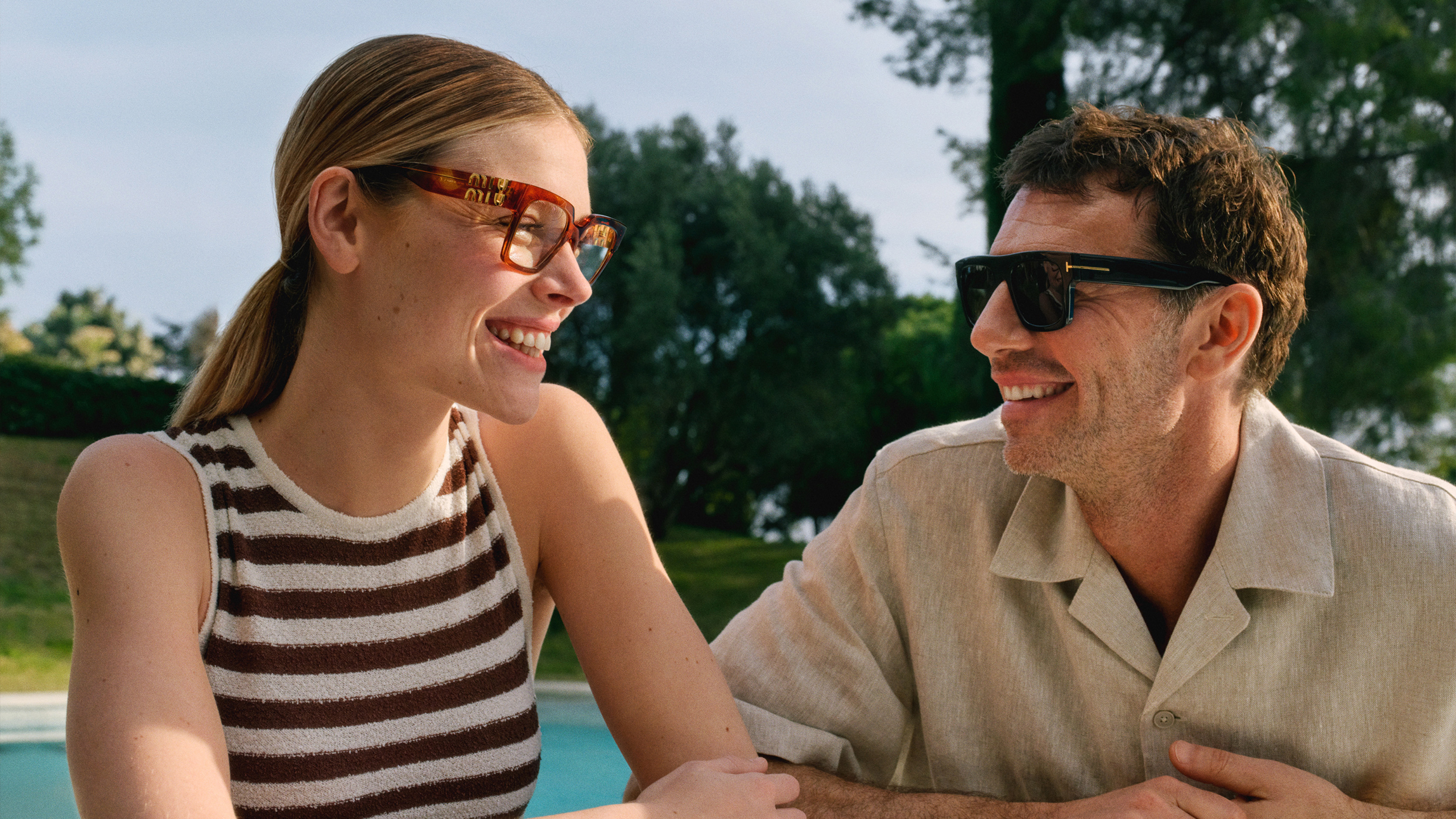 Un uomo e una donna con gli occhiali da sole sono seduti all'aperto vicino a una piscina, circondati da alberi, e si scambiano un sorriso.