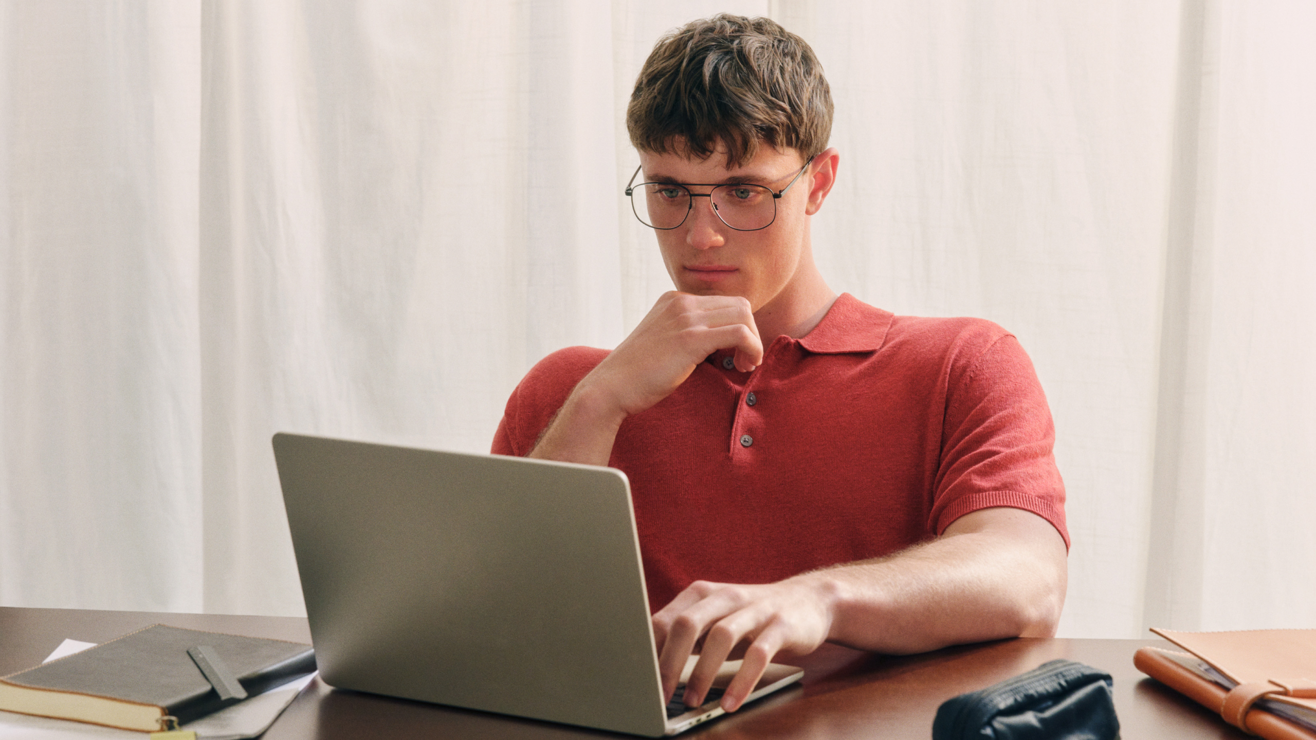 Person trägt eine Brille und ein rotes Hemd und blickt nachdenklich auf den Bildschirm eines Laptops. Die Person sitzt vor einem hellen Vorhang.