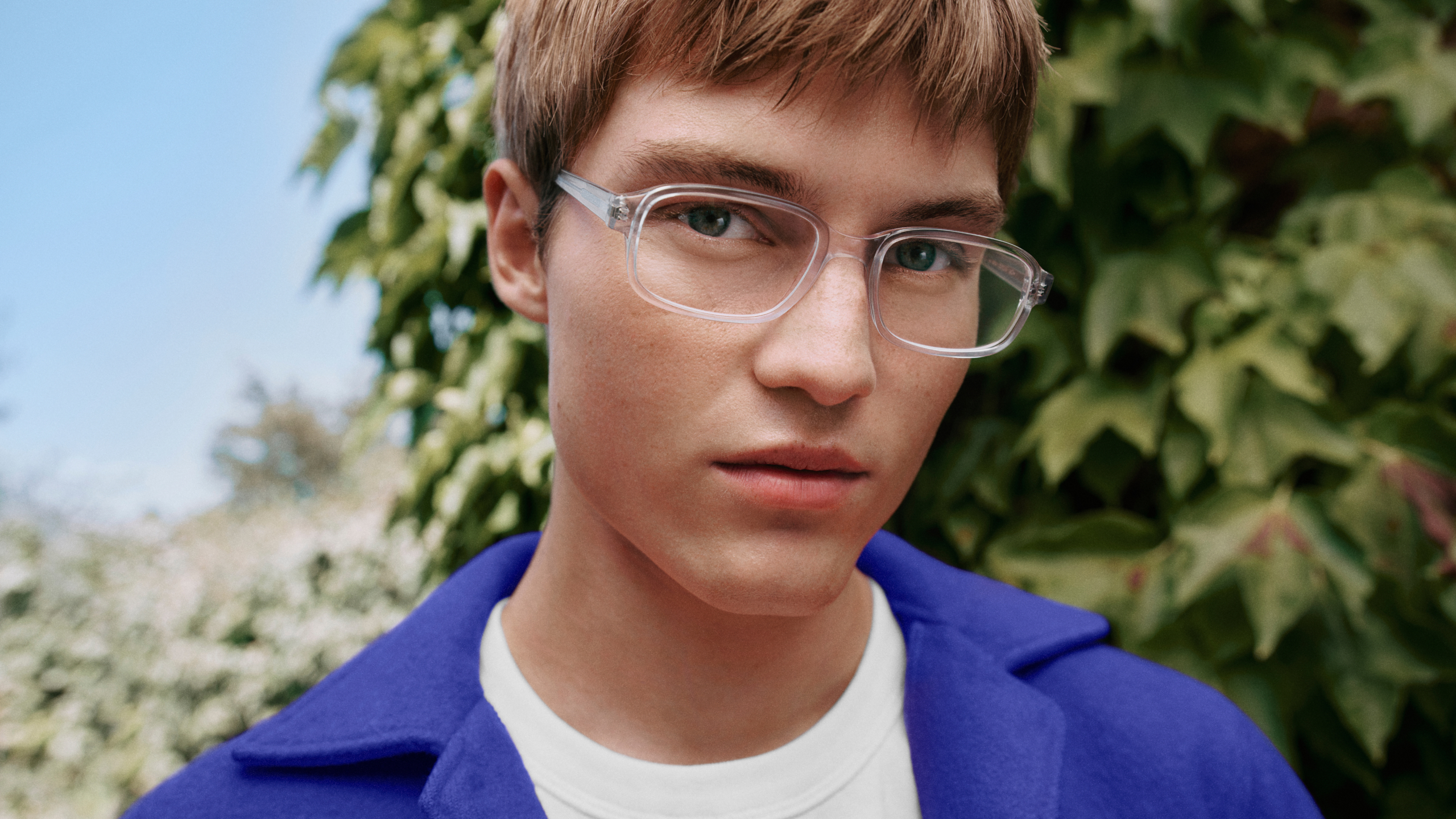 Eine Person mit transparenter Brille und blauer Jacke steht vor grünem Blattwerk, lächelt leicht und blickt in die Kamera.