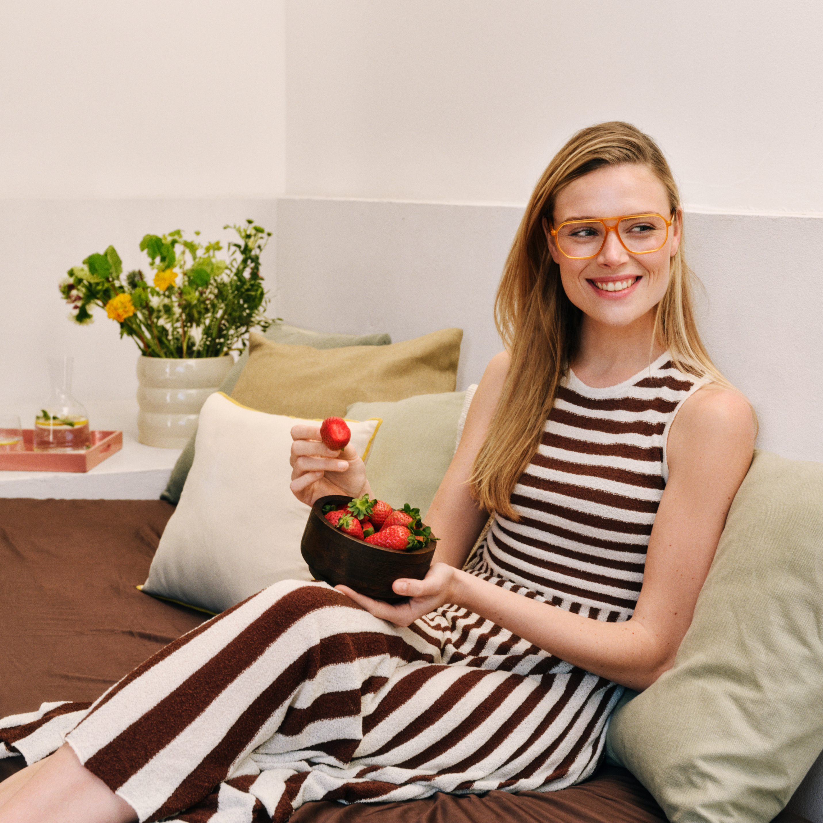 Frau in einem gestreiften Kleid isst Erdbeeren und sitzt auf einem Bett mit bunten Kissen. Im Hintergrund sind Blumen zu sehen.