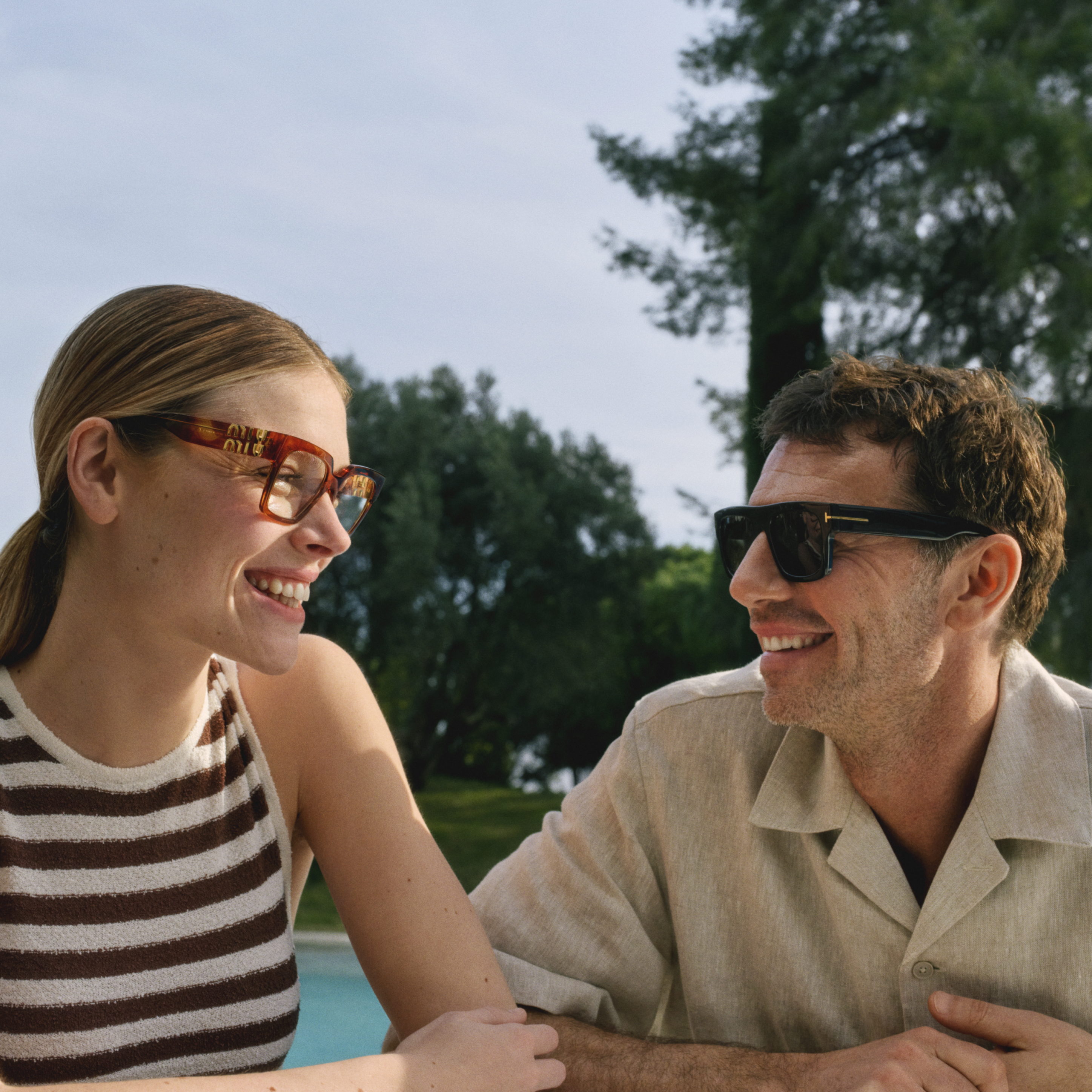 Zwei Menschen mit Sonnenbrillen lachen gemeinsam am Pool, umgeben von üppigem Grün an einem sonnigen Tag.