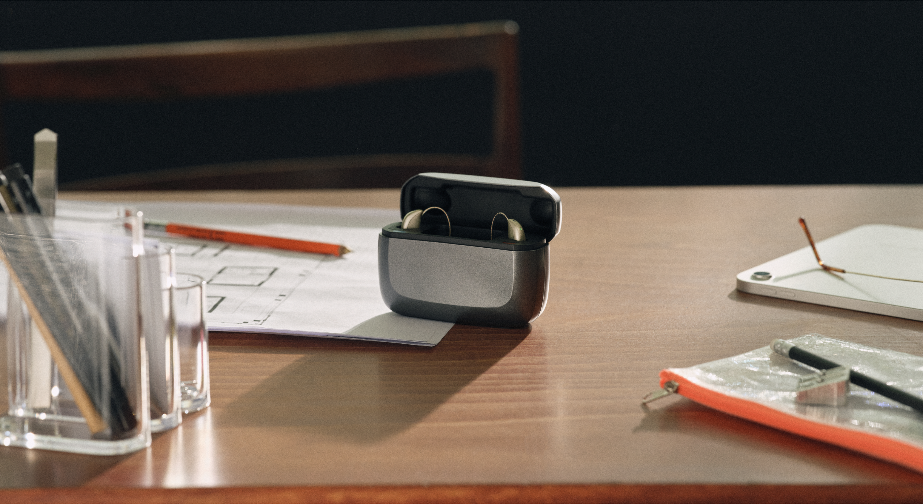 Hörgeräte in einem Ladecase auf einem Holzschreibtisch mit Papieren, einem Bleistift, einem Lineal in einem Halter und einer Reißverschlusstasche daneben.