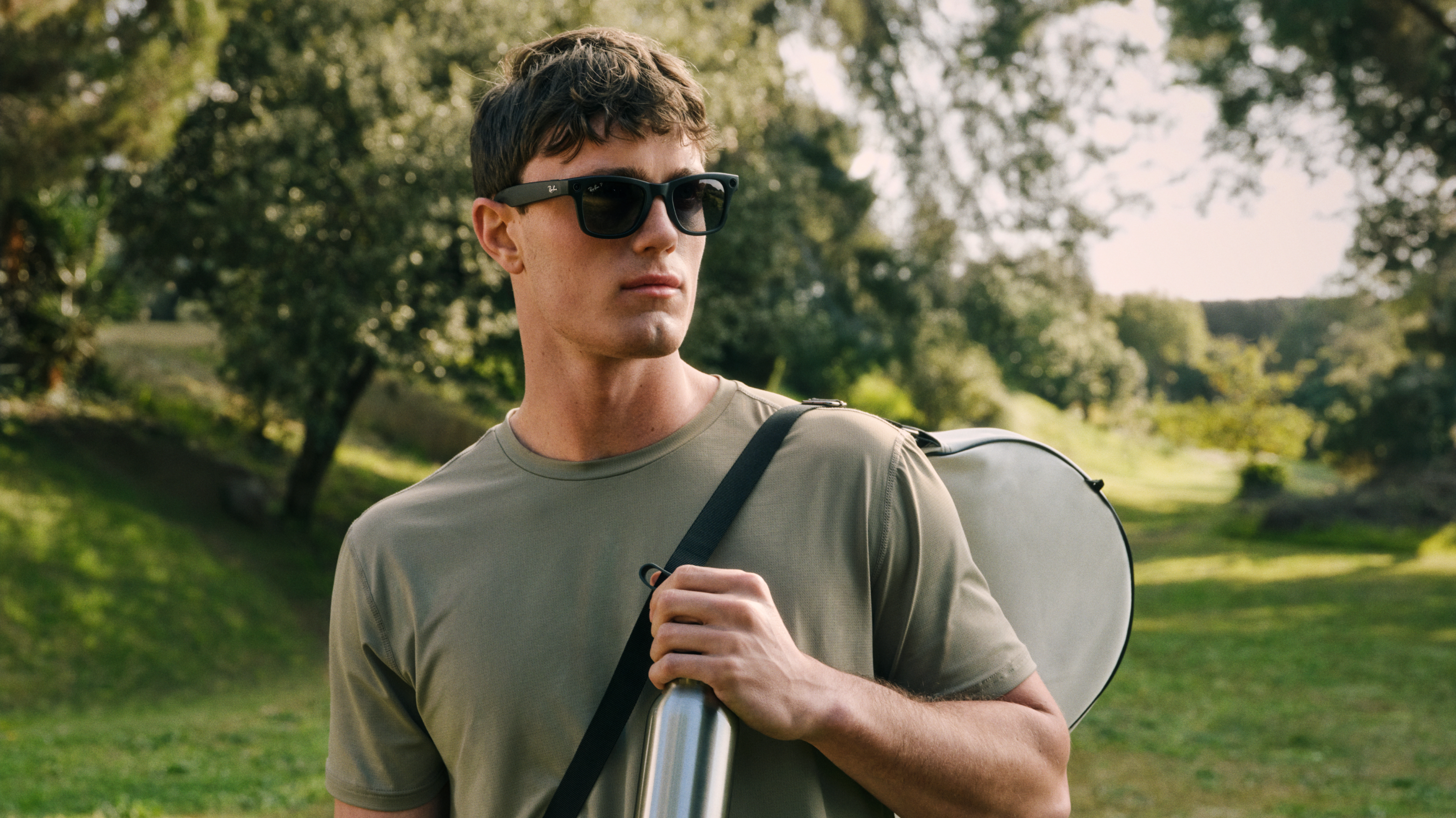 Ein Mann mit Sonnenbrille, der eine aufgerollte Yogamatte in der Hand hält, spaziert durch einen sonnigen Park, im Hintergrund sind Bäume und Gras zu sehen.