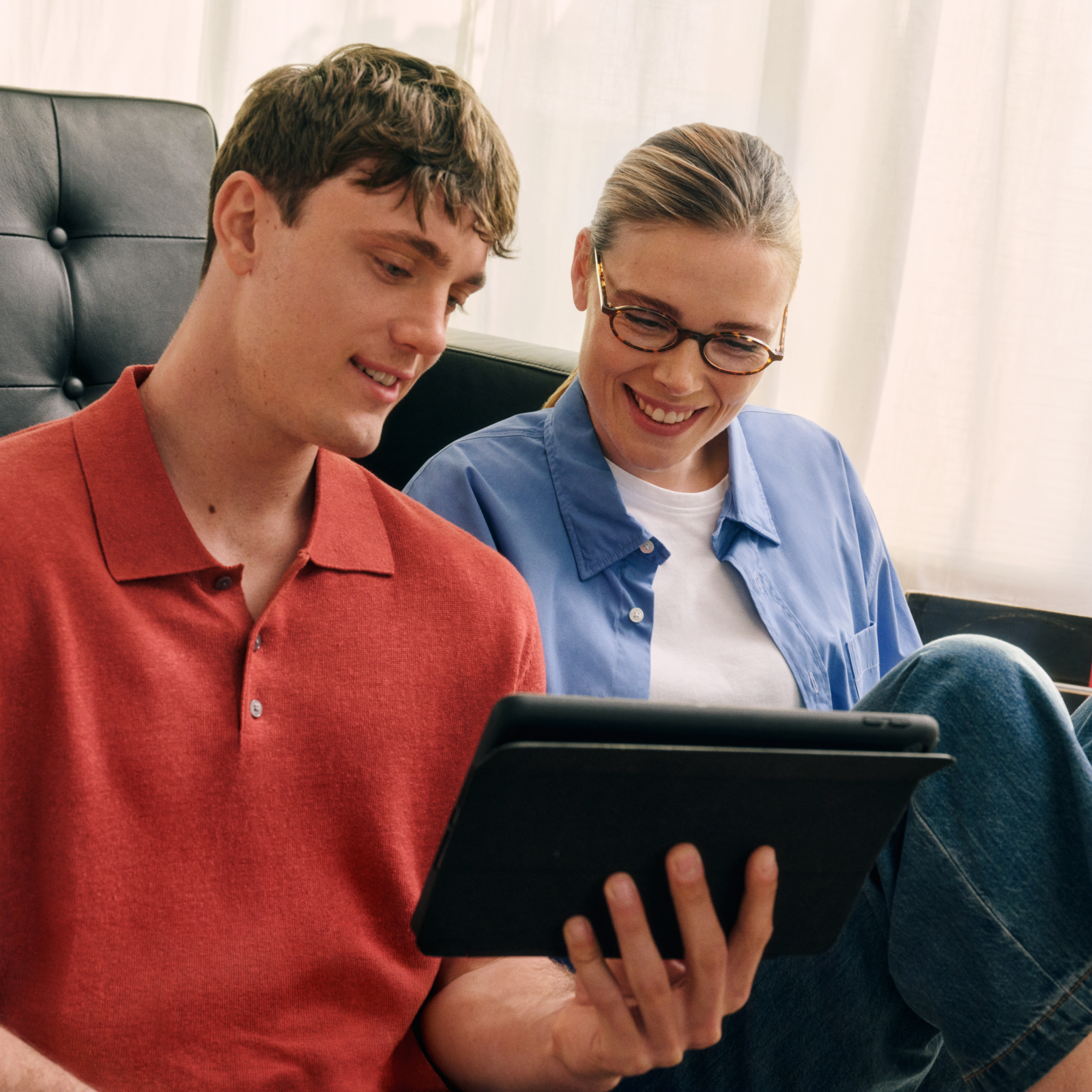 Un uomo e una donna sono seduti su un divano, sorridono e guardano insieme un tablet in una stanza ben illuminata.