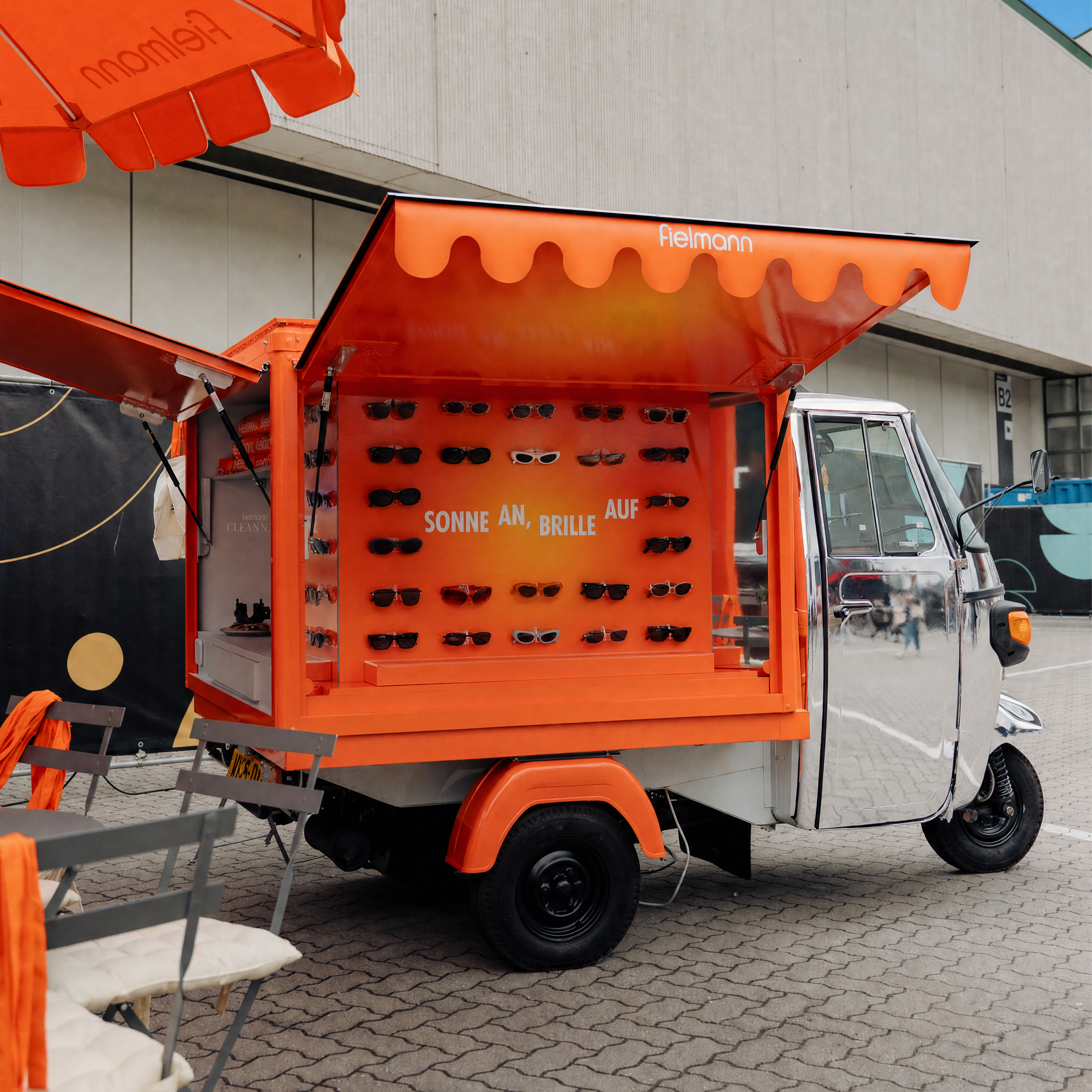 Ein orangefarbener Sonnenbrillenständer mit offenem Dach, auf dem Reihen von Sonnenbrillen ausgestellt sind und der Schriftzug „Sonne an, Brille auf“ zu sehen ist, steht in einer städtischen Straße.