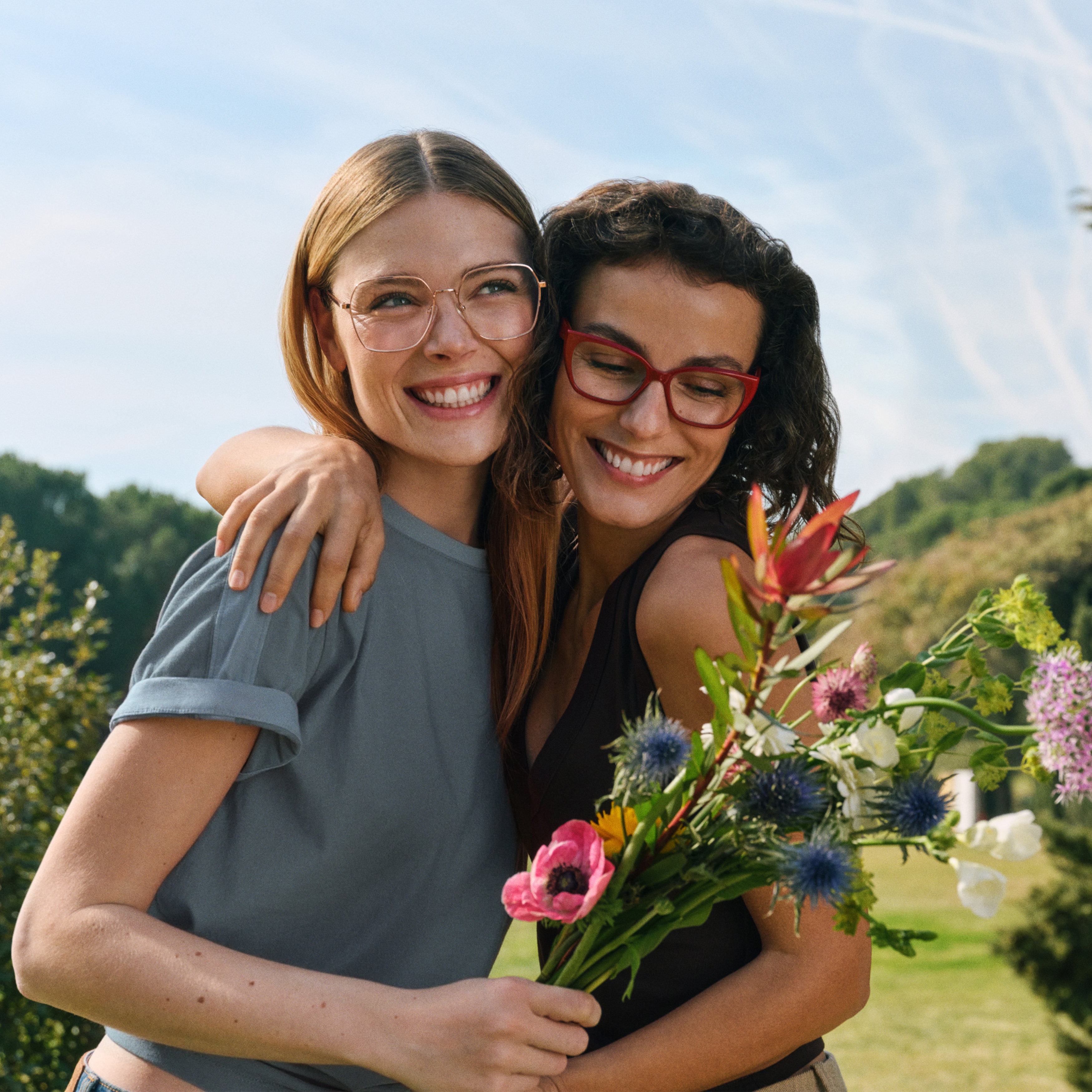 Due donne sorridenti con gli occhiali, una delle quali tiene in mano un mazzo di fiori colorati. Si abbracciano all'aperto; sullo sfondo si vedono piante verdi e un cielo azzurro.
