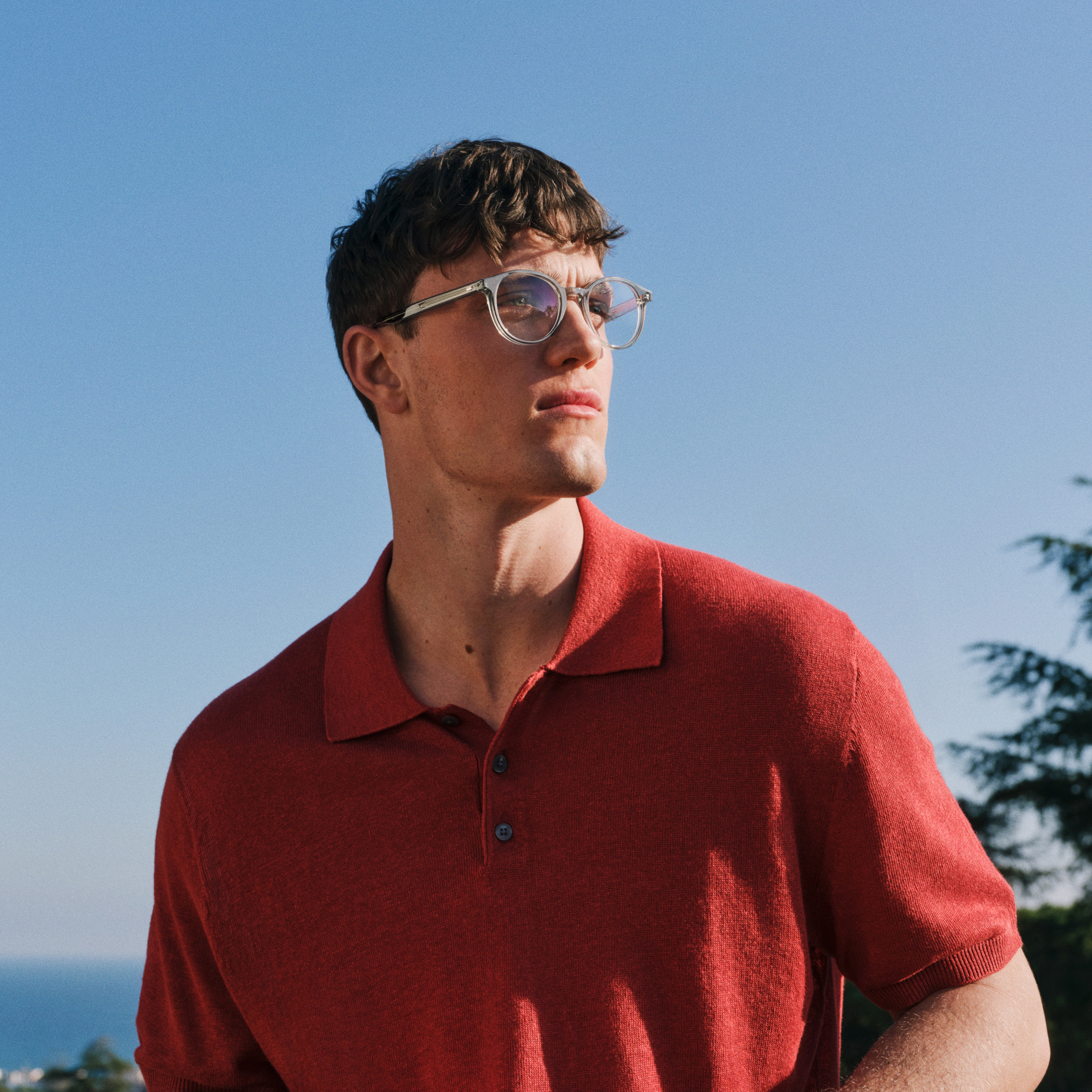 Un uomo con una polo rossa e occhiali chiari guarda di lato; sullo sfondo si vedono degli alberi e il cielo blu.