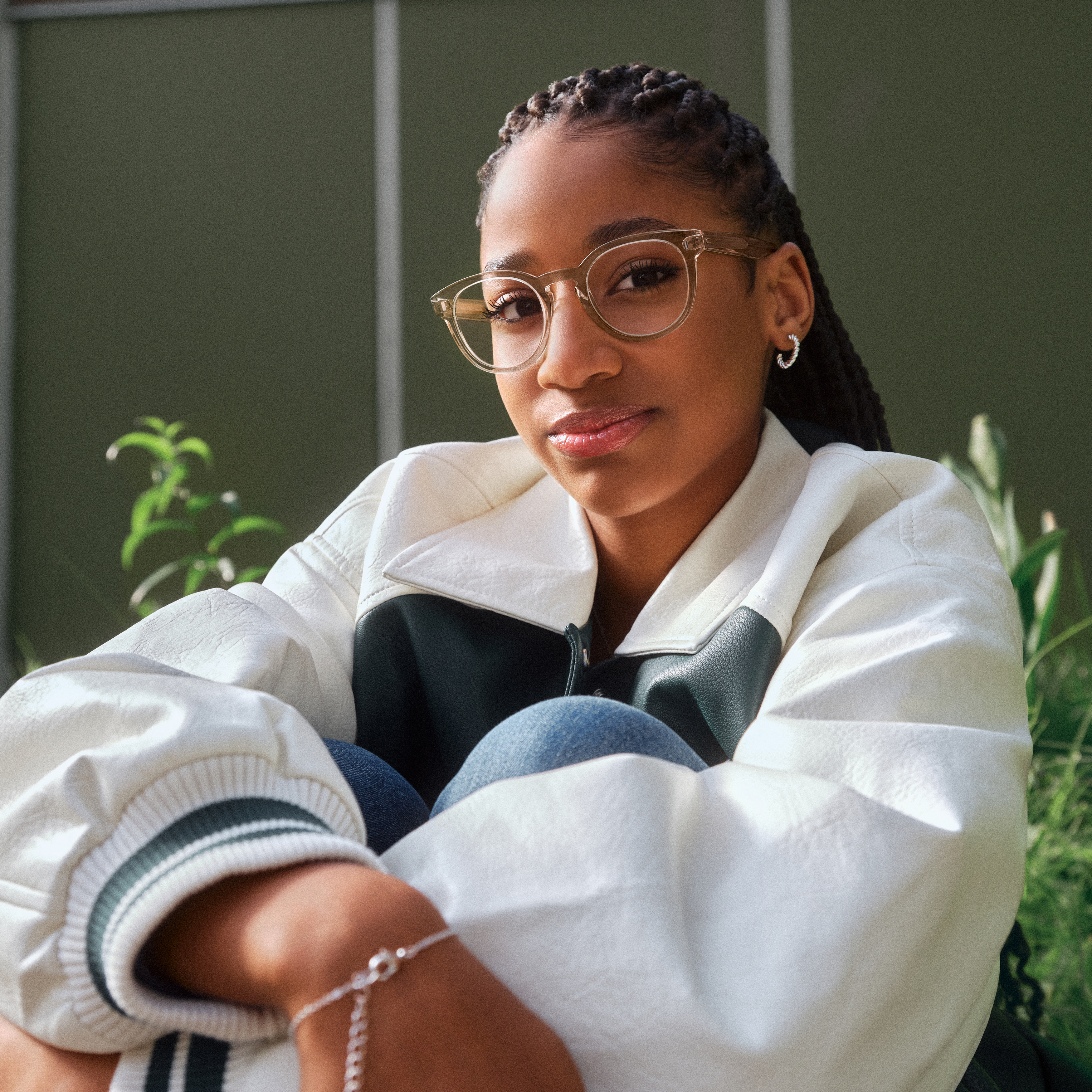 Eine Person mit Brille und weißer Jacke sitzt im Freien, umgeben von grünen Pflanzen, und blickt mit neutralem Gesichtsausdruck in die Kamera.