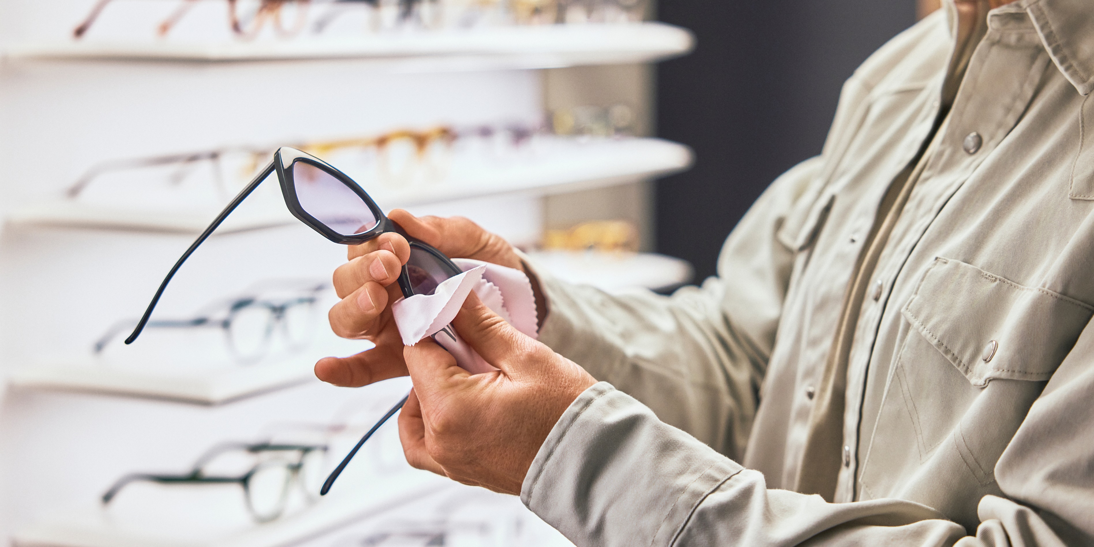 Eine Person reinigt in einem Brillengeschäft eine Brille mit einem Tuch, umgeben von Regalen mit verschiedenen Brillenfassungen.