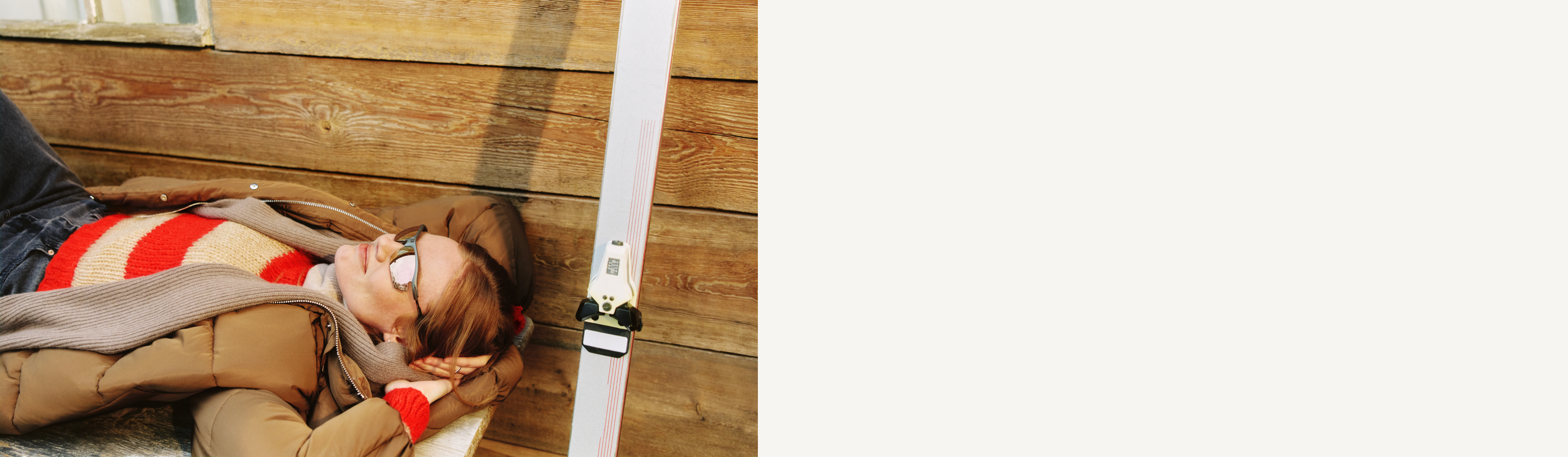 Person in einem rot-beige gestreiften Pullover und einer braunen Jacke, die auf einer Holzbank liegt, neben sich die Skier abgestützt, und einen sonnigen Tag genießt.