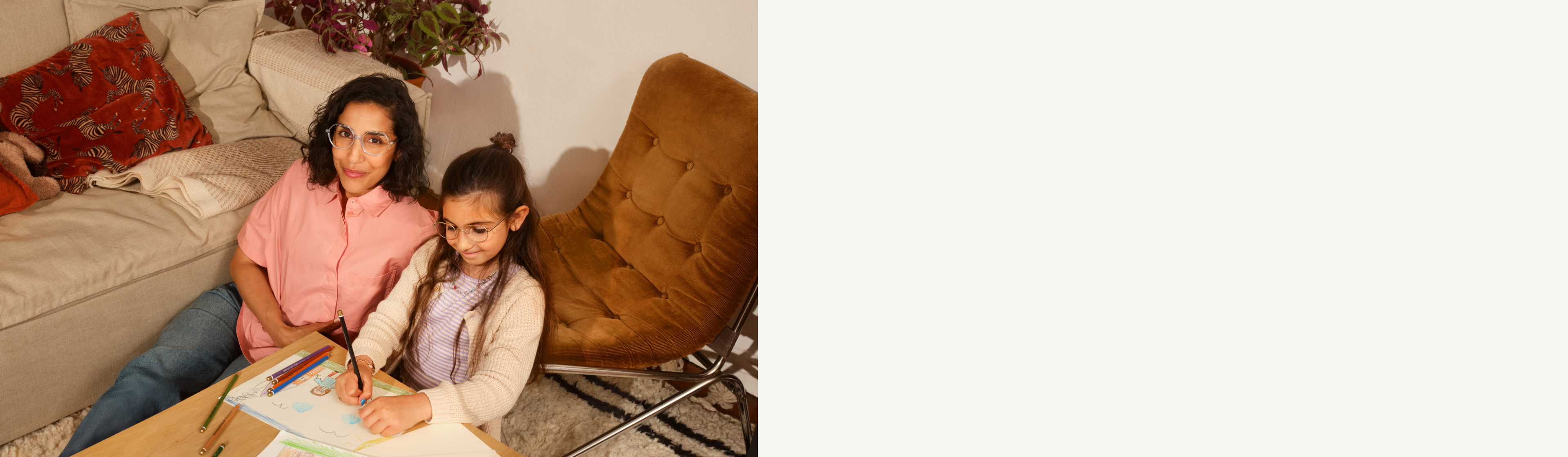 Eine Frau und ein Mädchen zeichnen gemeinsam an einem kleinen Tisch, umgeben von Buntstiften, in einer gemütlichen Wohnzimmeratmosphäre.