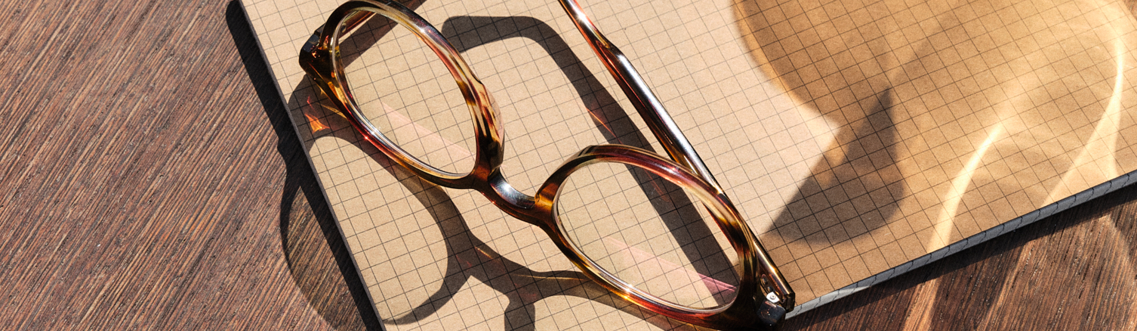 Brille in Schildpatt-Optik liegt auf einem geöffneten karierten Notizbuch und wirft Schatten auf eine sonnenbeschienene Holzoberfläche.