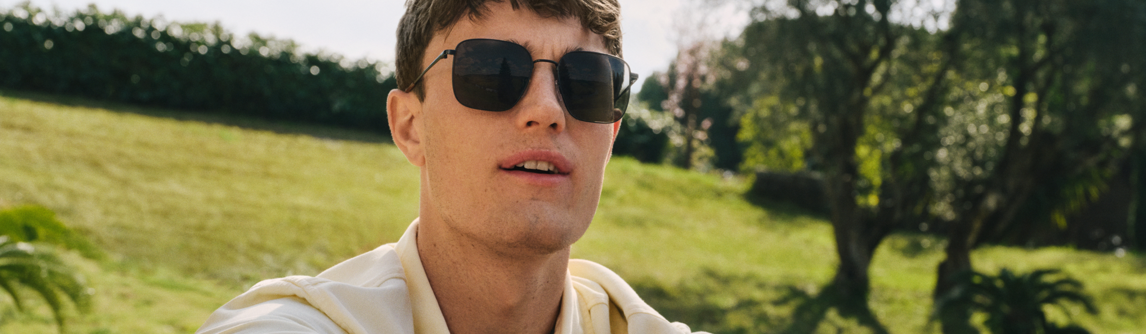 Eine Person mit Sonnenbrille und hellem Hemd sitzt an einem sonnigen Tag im Freien auf dem Rasen, im Hintergrund sind Bäume und Büsche zu sehen.