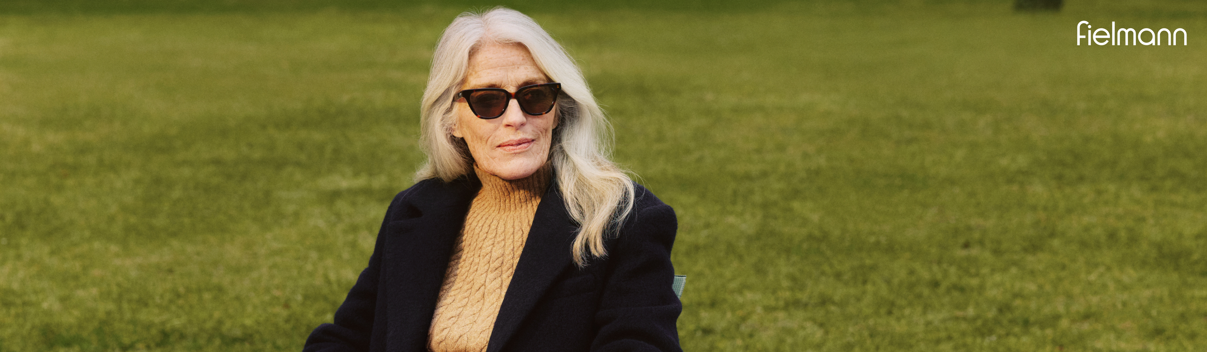 Starsza kobieta o długich białych włosach, w okularach przeciwsłonecznych i ciemnym płaszczu siedzi na ławce w parku, w rogu widnieje napis „Fielmann”.