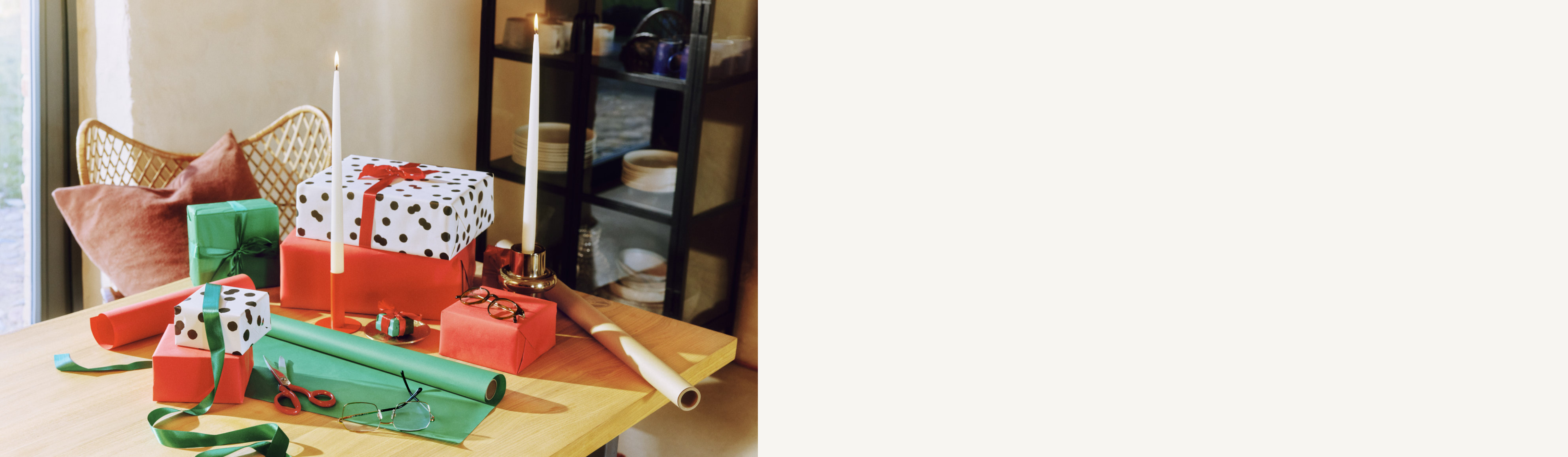 Holztisch mit zwei Brillen sowie eingepackten Geschenken in grünem, rotem und gepunktetem Papier, dazu Bänder, eine Schere und Geschenkpapierrollen neben einem Stuhl.