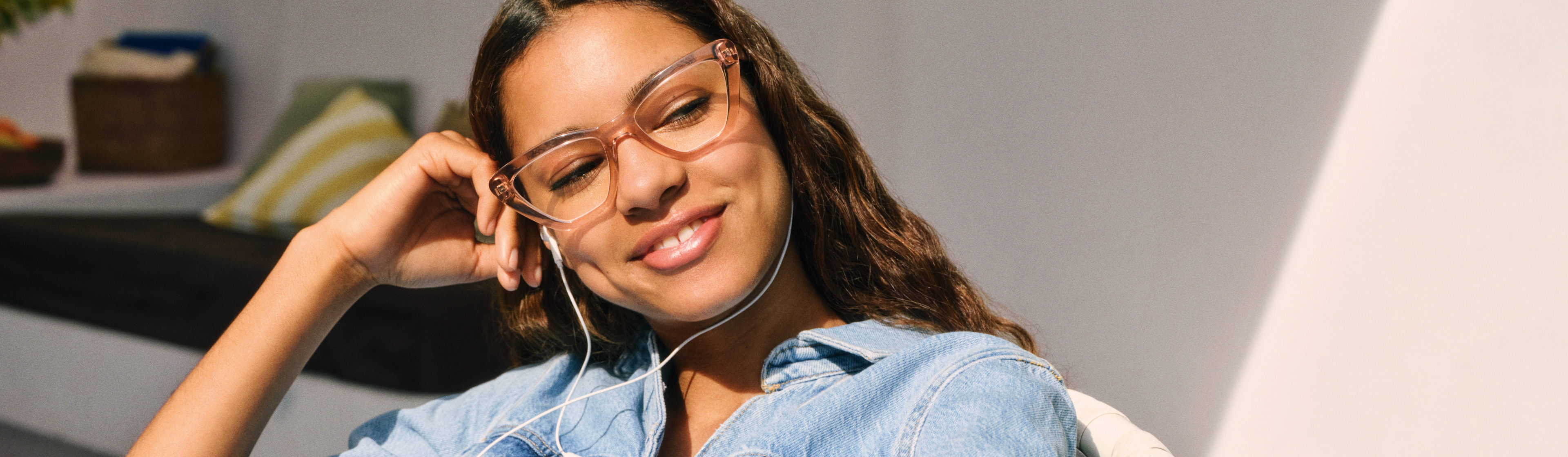Donna con giacca di jeans e occhiali, sorridente, con auricolari, seduta in modo rilassato, con la testa appoggiata sulla mano.