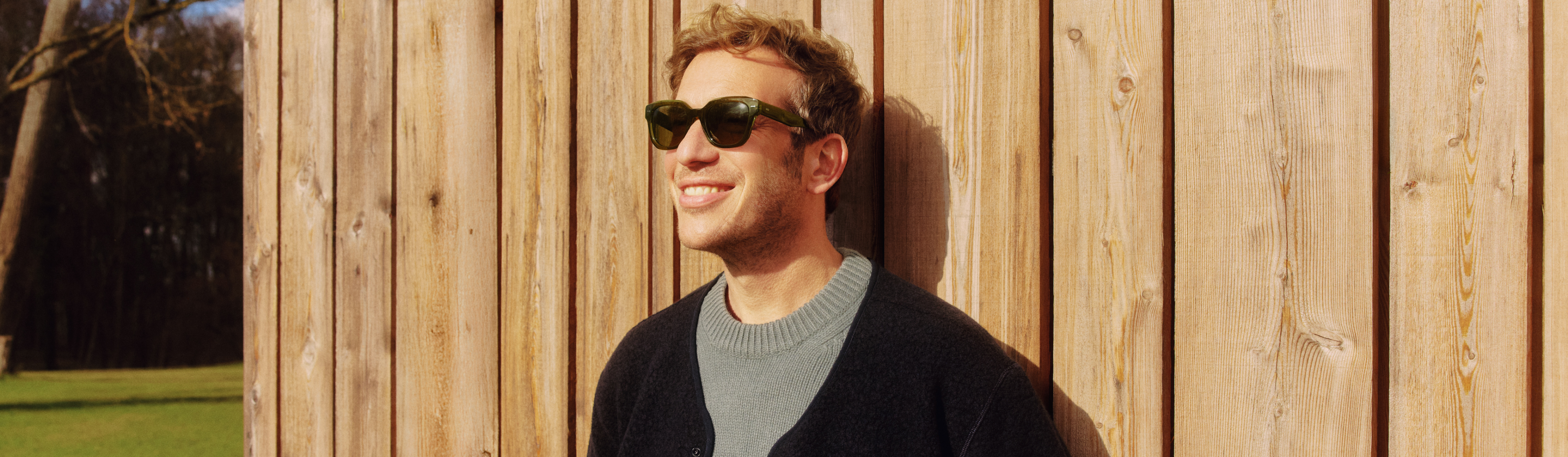 Ein Mann mit Sonnenbrille steht lächelnd vor einer Holzwand und genießt die Sonne. Er trägt einen grauen Pullover und eine schwarze Jacke.