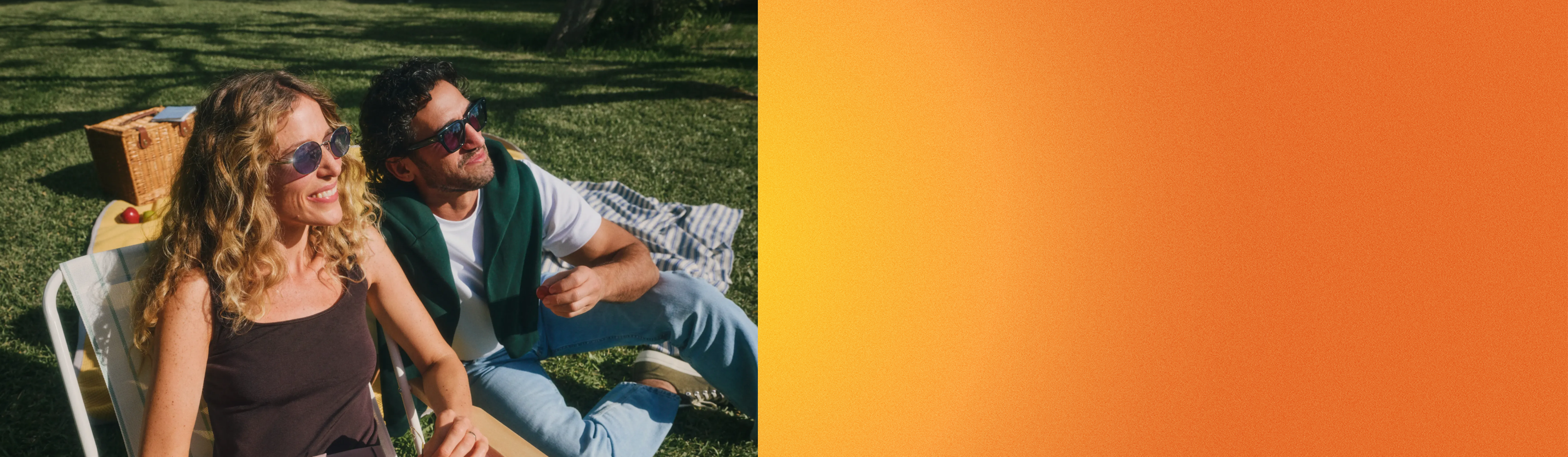 Ein Paar mit Sonnenbrillen entspannt sich an einem sonnigen Tag im Freien, sitzt auf Gartenstühlen und hat einen Picknickkorb neben sich stehen.