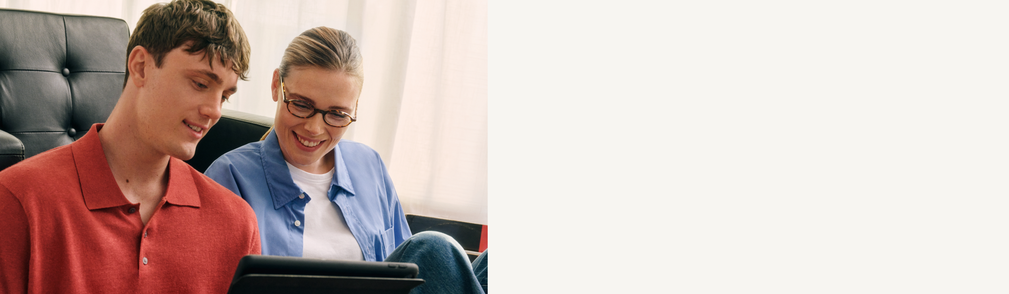 Ein Mann und eine Frau sitzen auf einem Sofa, lächeln und schauen gemeinsam auf ein Tablet in einem gut beleuchteten Raum.