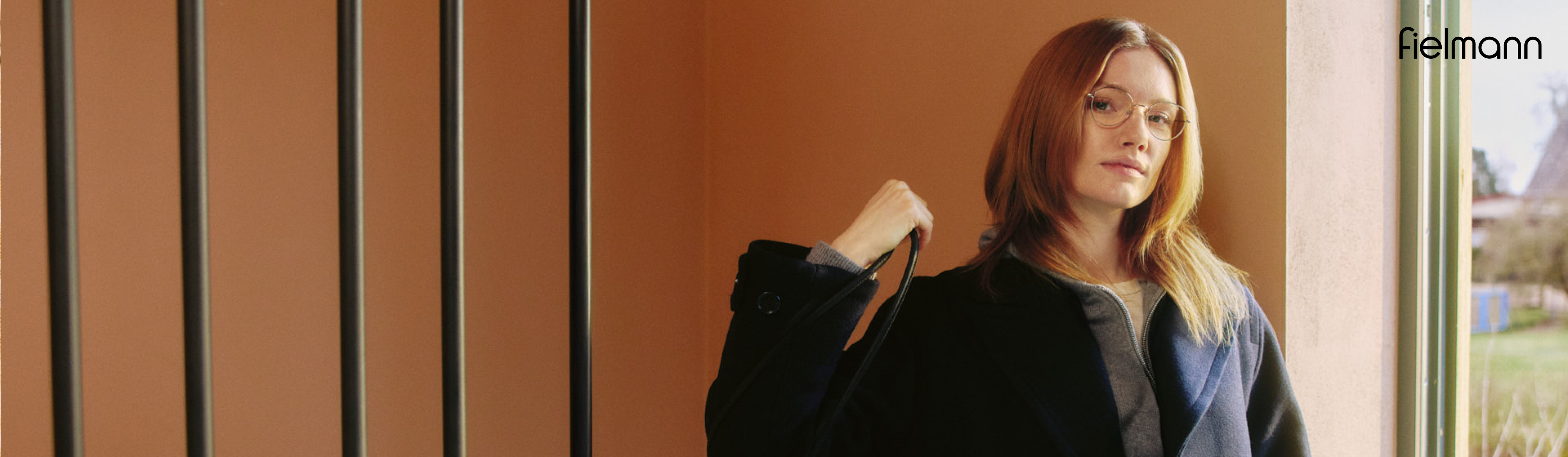 Kobieta w okularach, ubrana w ciemny płaszcz, pozuje w pomieszczeniu przed pomarańczową ścianą, trzymając w dłoni pasek do torebki.