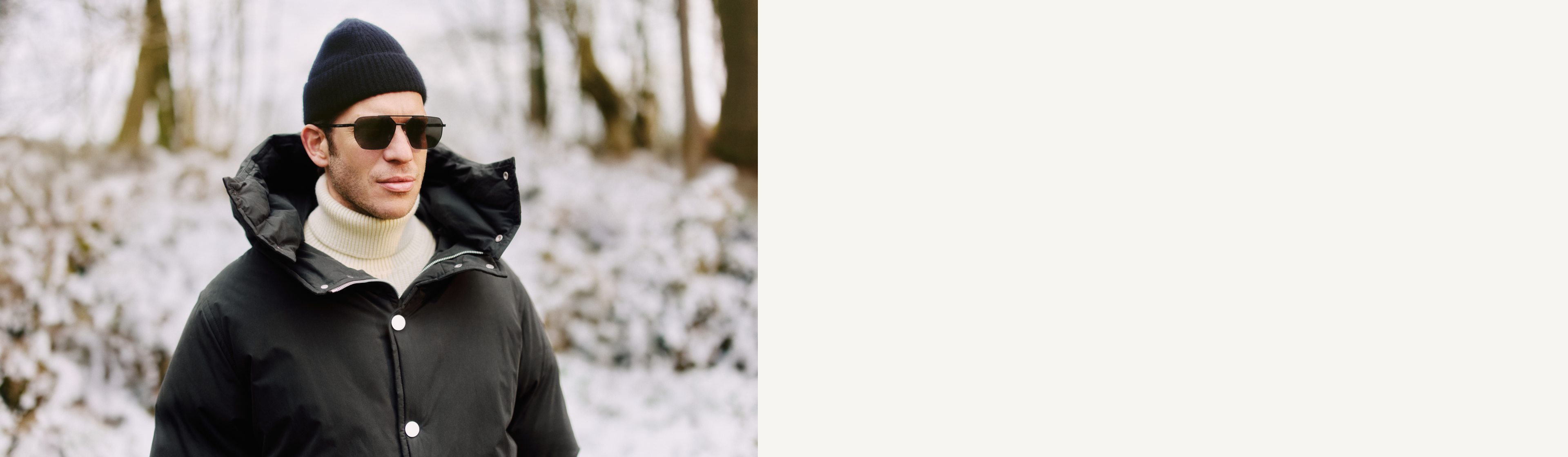 Uomo con cappotto invernale nero, cappello e occhiali da sole in piedi all'aperto in un ambiente innevato, lo sguardo rivolto di lato.