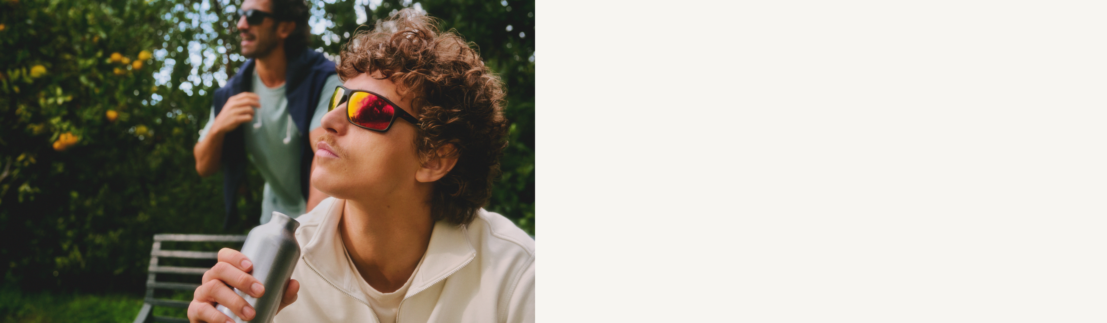 Zwei Herren im Freien mit Sonnenbrillen; einer trinkt aus einer Flasche. Im Hintergrund befinden sich üppiges Grün und eine Bank.