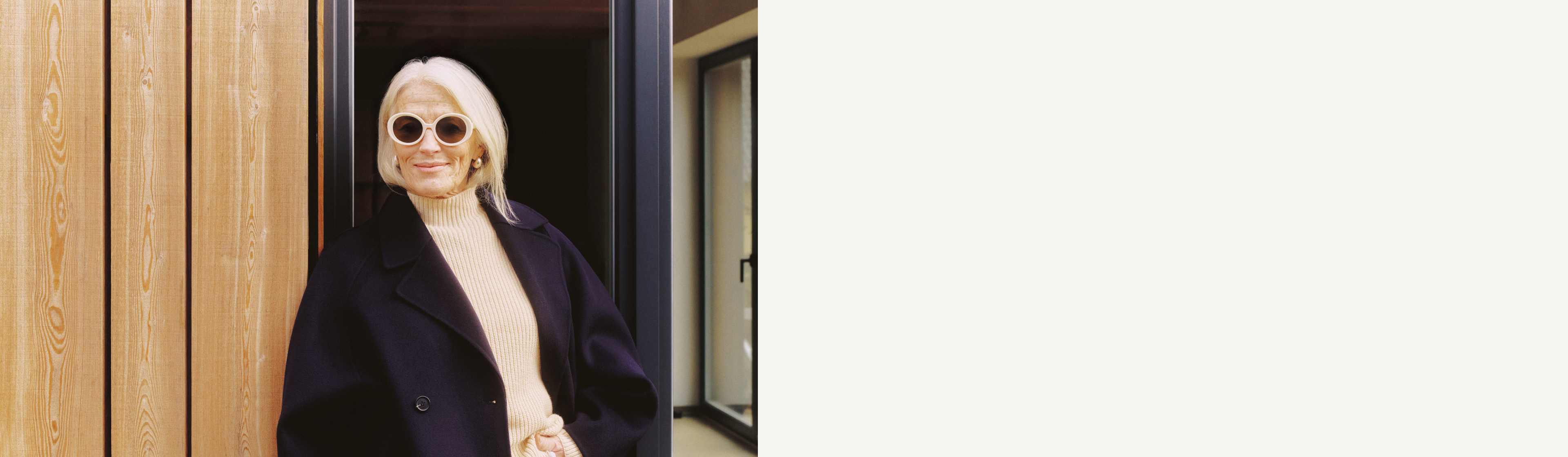 Ältere Frau mit weißem Haar, runder Sonnenbrille, beigem Rollkragenpullover und schwarzem Mantel steht neben einer Glastür und einer Holzwand.