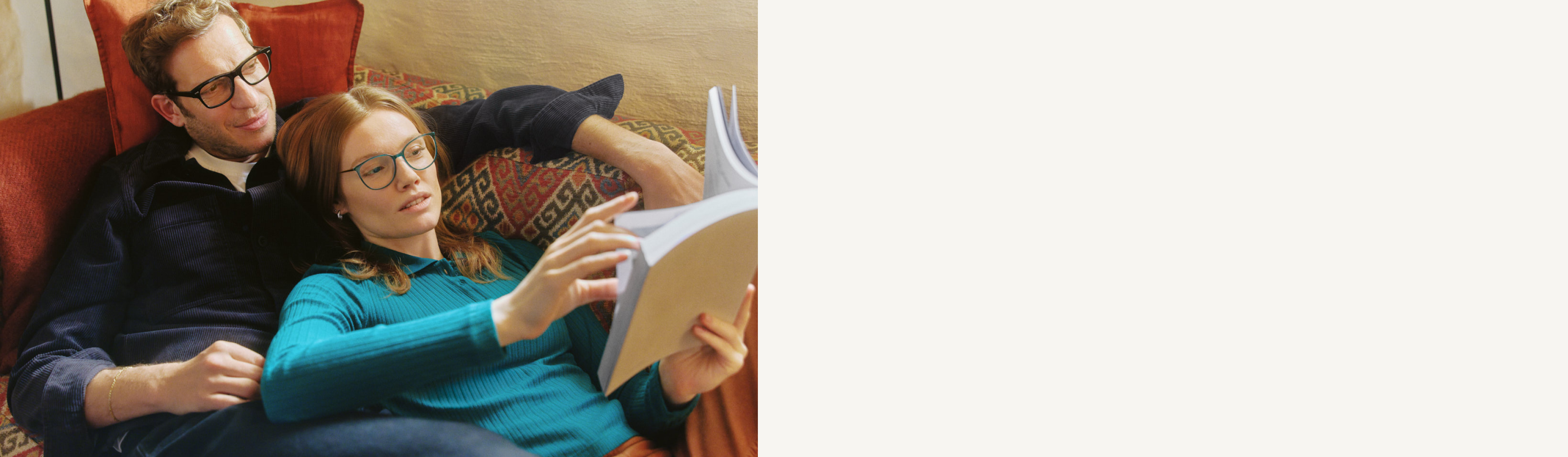 Ein Paar mit Brillen entspannt gemeinsam auf einem Sofa mit bunten Kissen und wirkt ruhig und zufrieden.