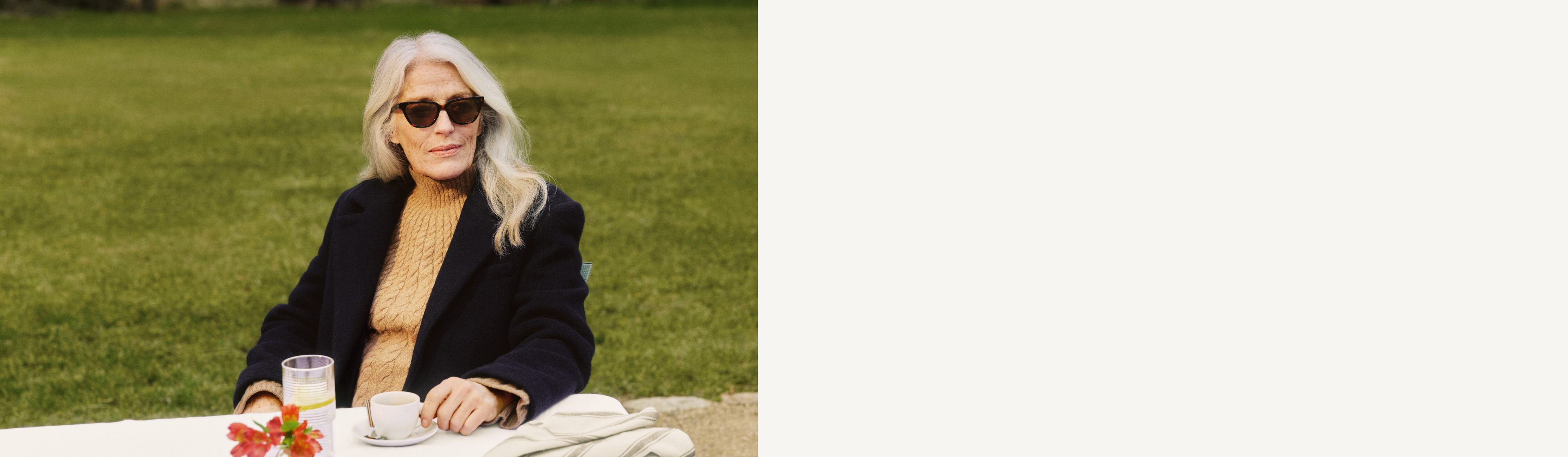 Žena s dlouhými vlasy a slunečními brýlemi sedí u venkovního stolu s nápojem a květinami.