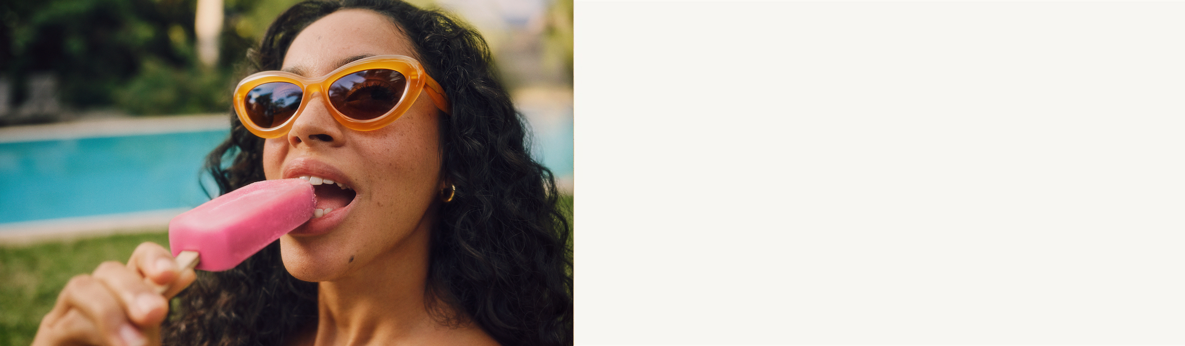 Una donna con occhiali da sole arancioni che si gode un ghiacciolo rosa, con una piscina sfocata sullo sfondo.