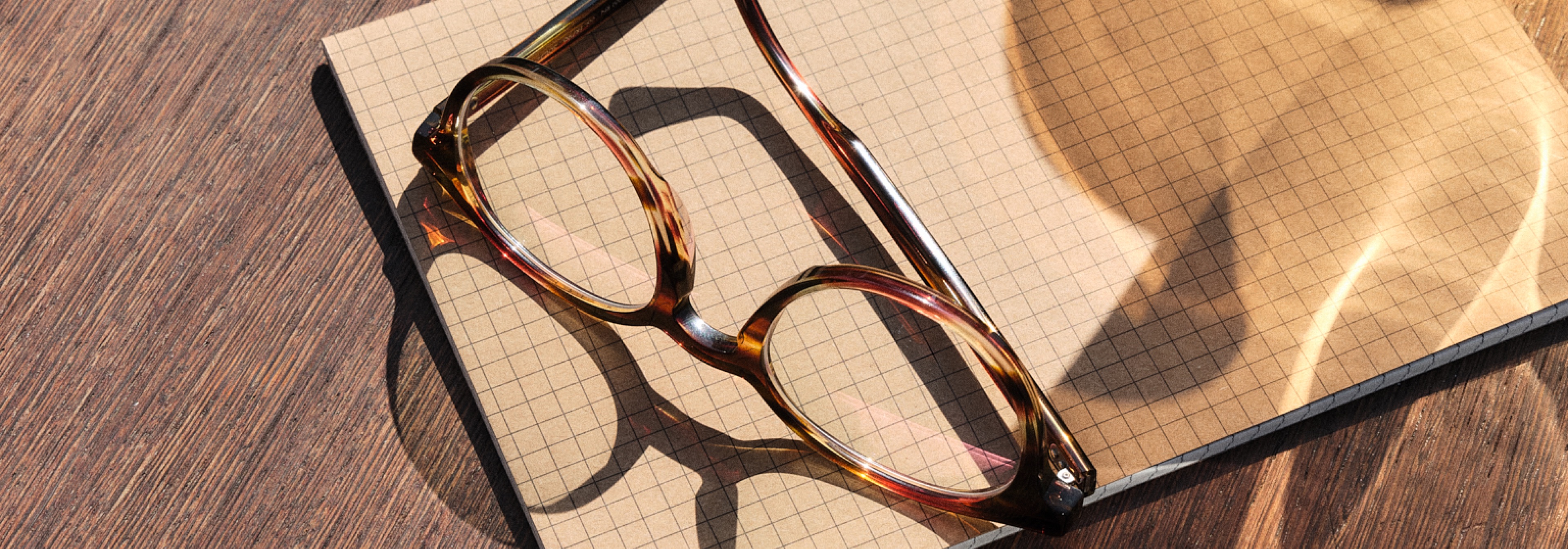 Brille in Schildpatt-Optik liegt auf einem geöffneten karierten Notizbuch und wirft Schatten auf eine sonnenbeschienene Holzoberfläche.