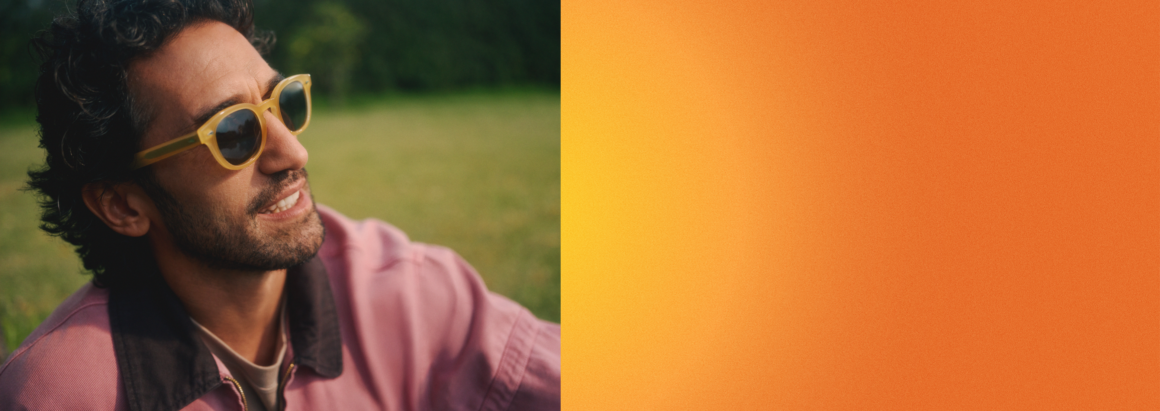 Un homme portant des lunettes de soleil et une veste violette est assis à l'extérieur, le regard pensif tourné vers le ciel, avec de l'herbe verte en arrière-plan.