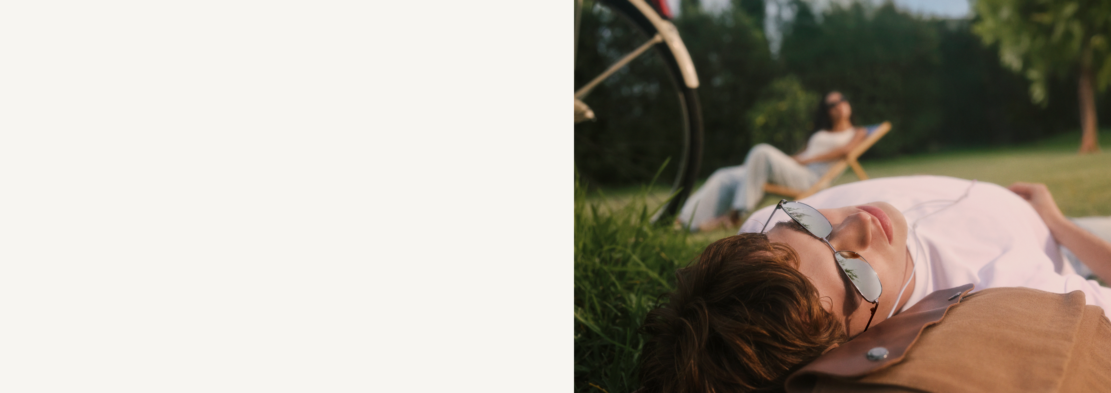 Person liegt mit Sonnenbrille und Kopfhörern auf dem Rasen, ein Fahrrad steht daneben. Eine weitere Person sitzt im Hintergrund auf einem Liegestuhl. Es ist ein strahlender, sonniger Tag.