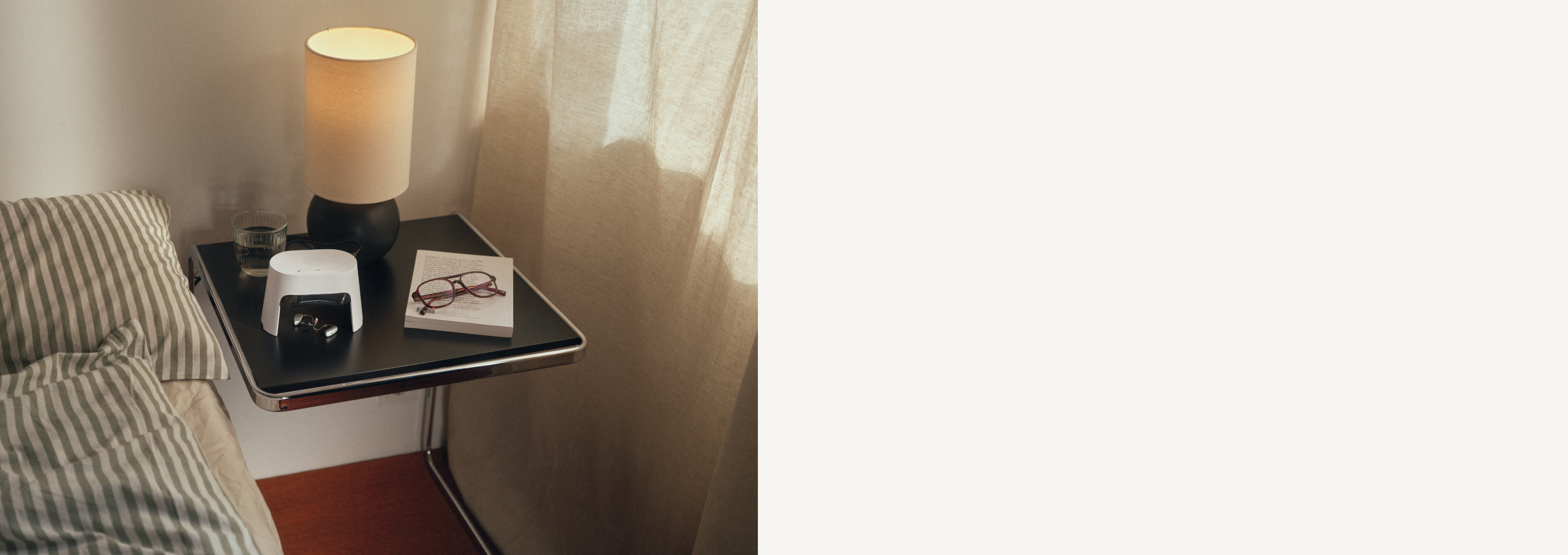 Ein gemütlicher Nachttisch mit einer Lampe, einer Brille, einem Buch und einem kleinen Gerät. Ein gestreiftes Kissen ist teilweise auf dem nahegelegenen Bett sichtbar.