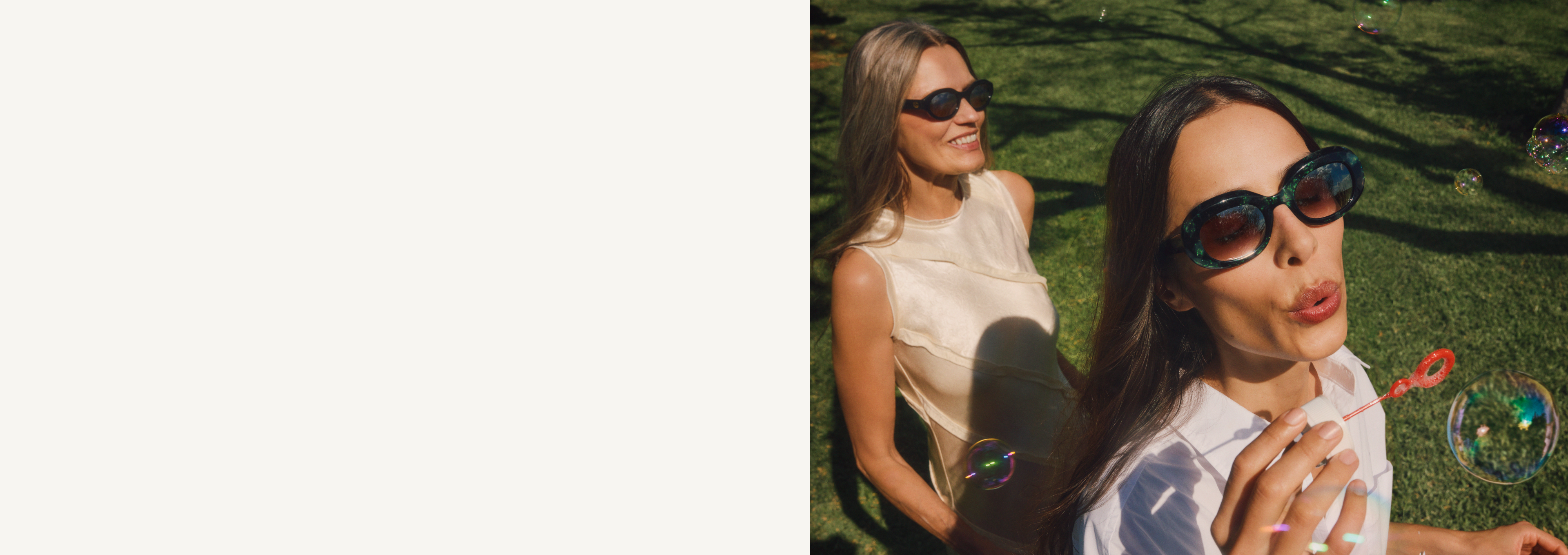 Zwei Frauen mit Sonnenbrillen genießen einen sonnigen Tag auf dem Rasen. Eine von ihnen bläst Seifenblasen und verleiht der Szene eine verspielte Note.