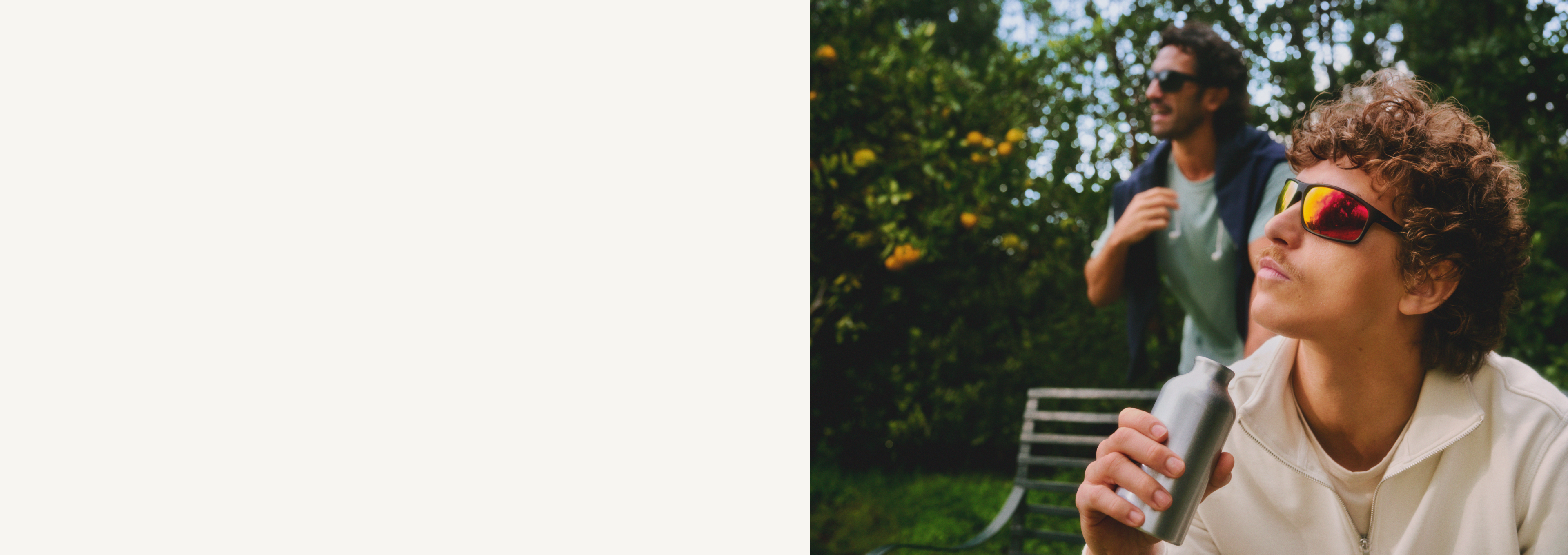 Zwei Personen im Freien; eine mit lockigem Haar und Sonnenbrille, die aus einer Flasche trinkt, die andere im Hintergrund mit Sonnenbrille und Jacke.