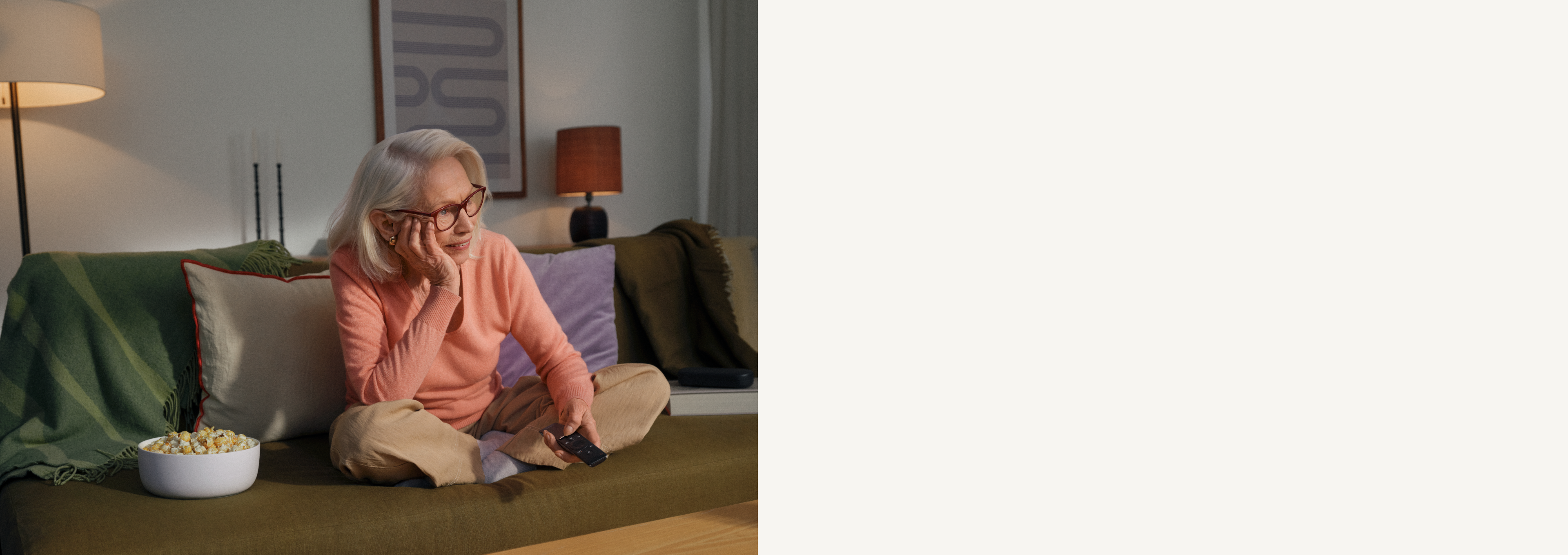 Une femme âgée portant des lunettes est assise sur un canapé et tient une télécommande dans la main.