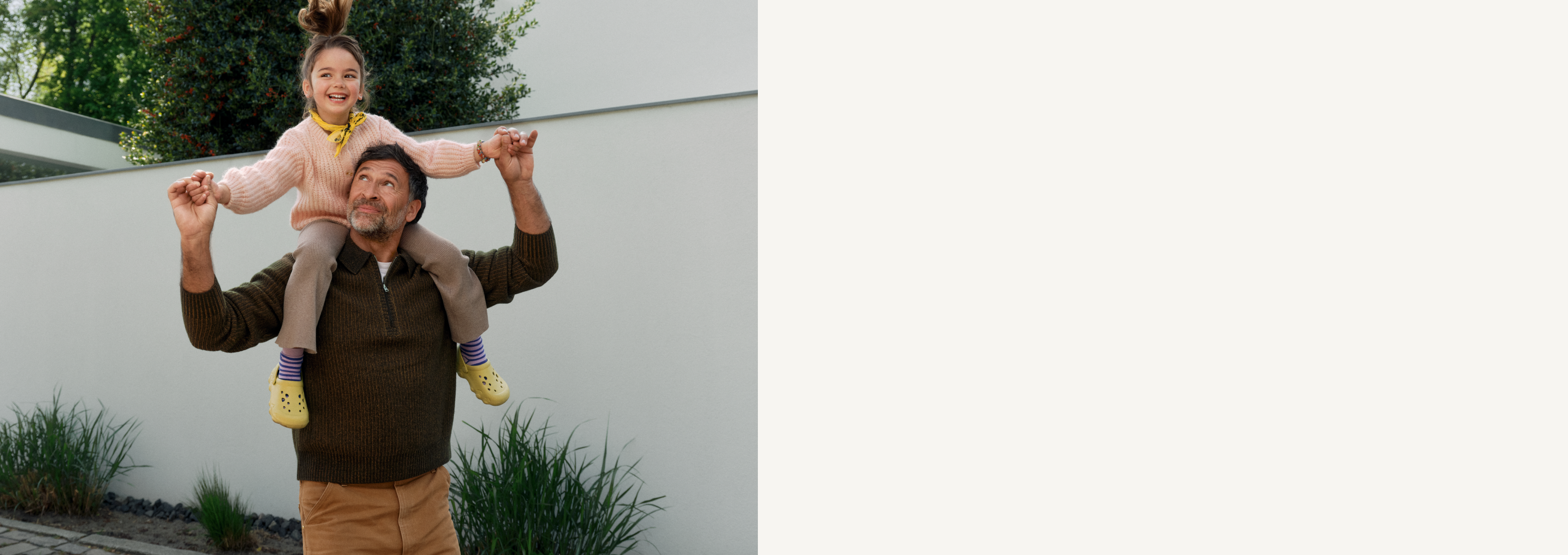 Ein Mann trägt ein lachendes Kind huckepack im Garten. Beide halten sich an den Händen und schauen nach oben.